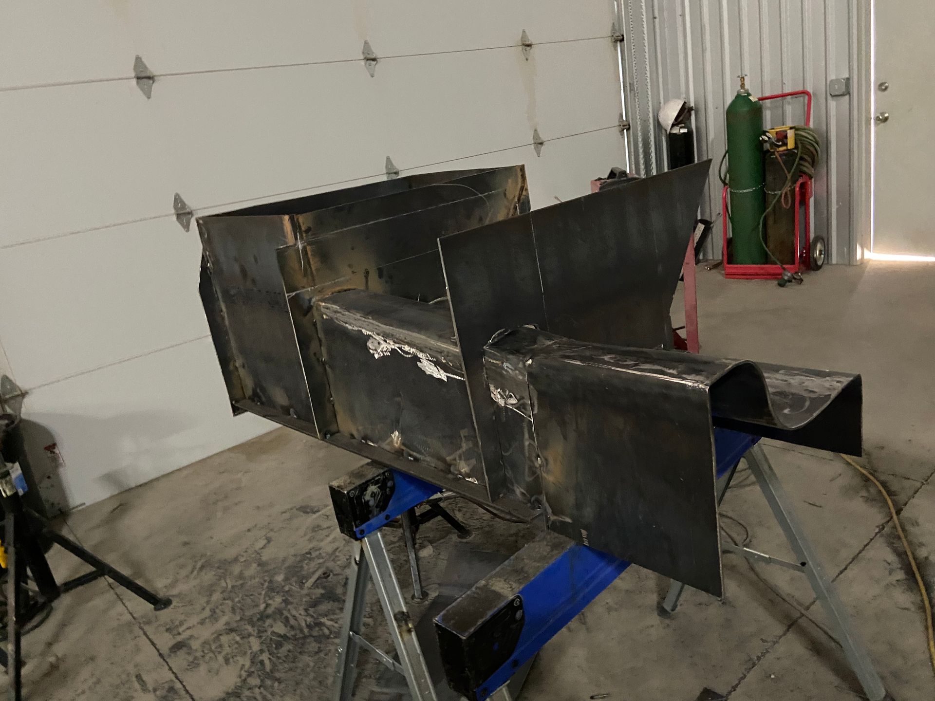 Welded steel fabrication, a dark box-like structure, resting on a blue sawhorse in a workshop.