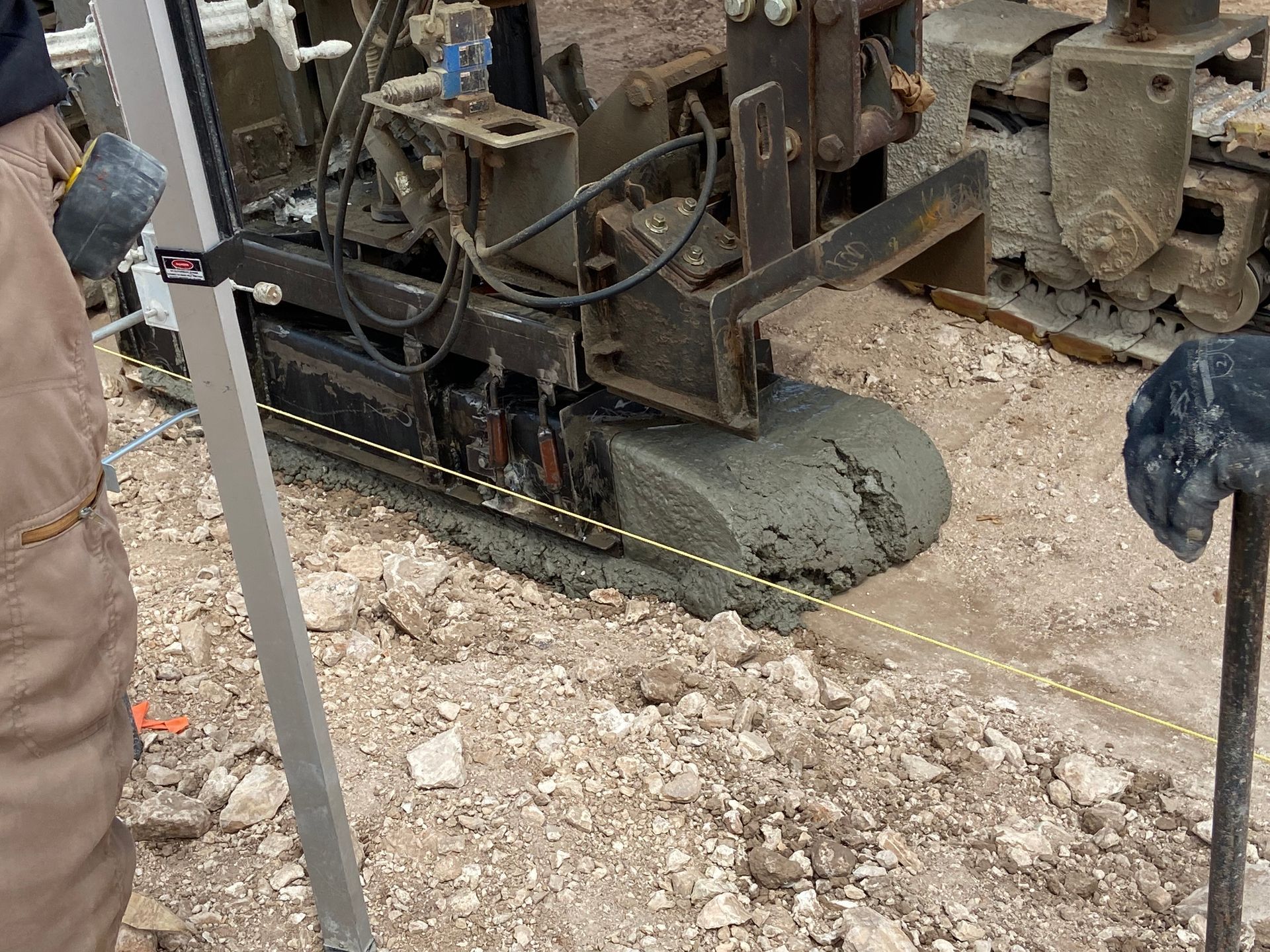 Construction equipment on gravel, pouring wet cement; a leveling rod is in the foreground.