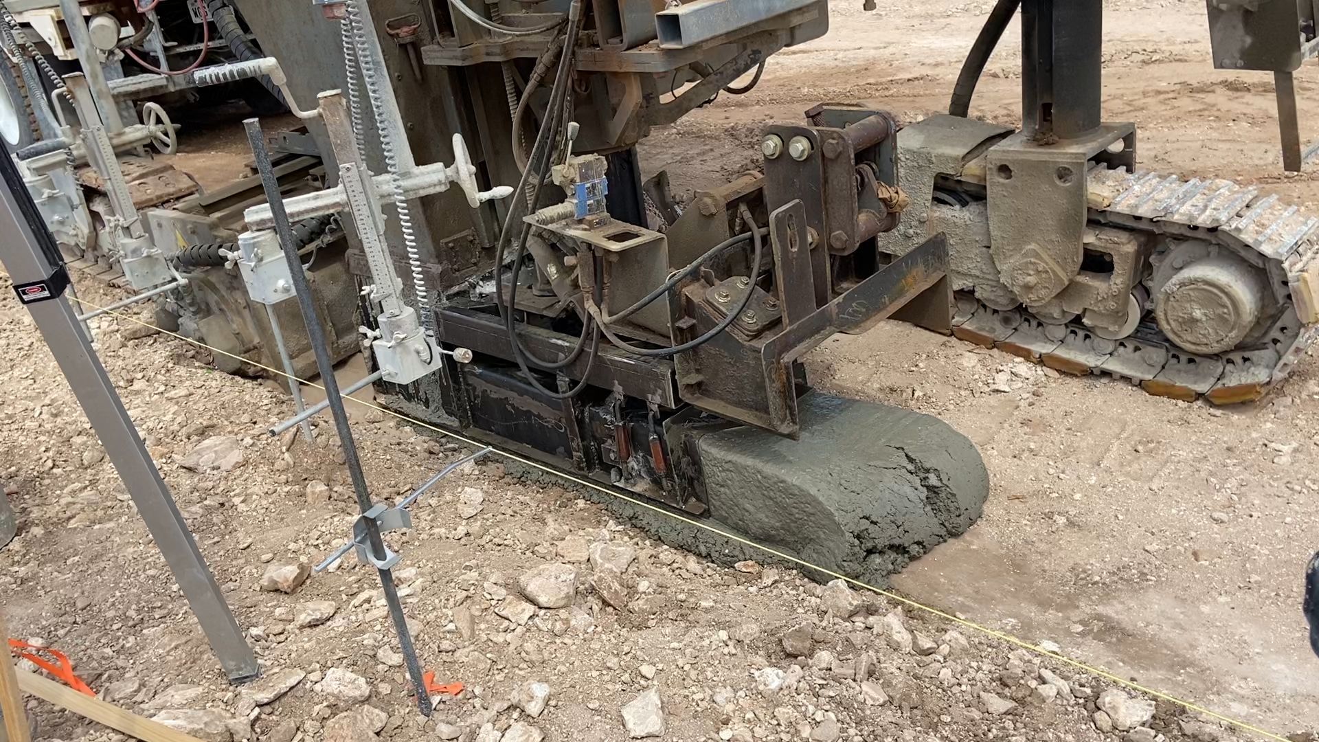 Concrete paving machine laying a strip of concrete on a dirt surface outdoors.