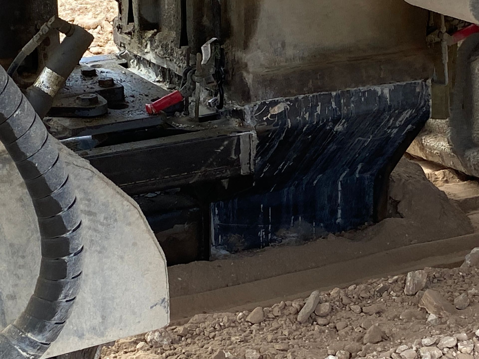 Black machinery component sitting on dusty ground, with hydraulic hoses visible.