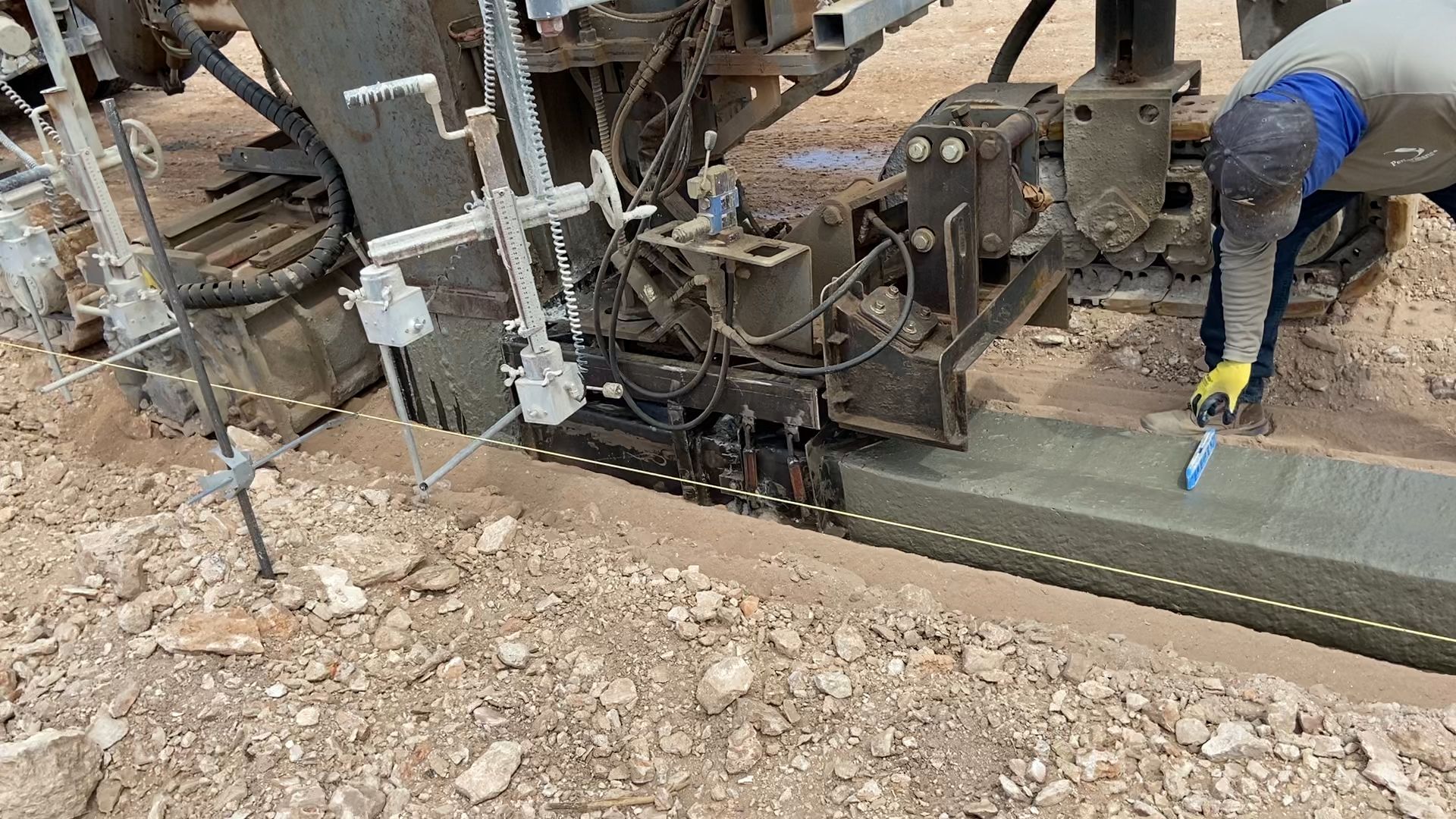 Construction worker smoothing concrete curb with a tool, machine in background, outdoors.