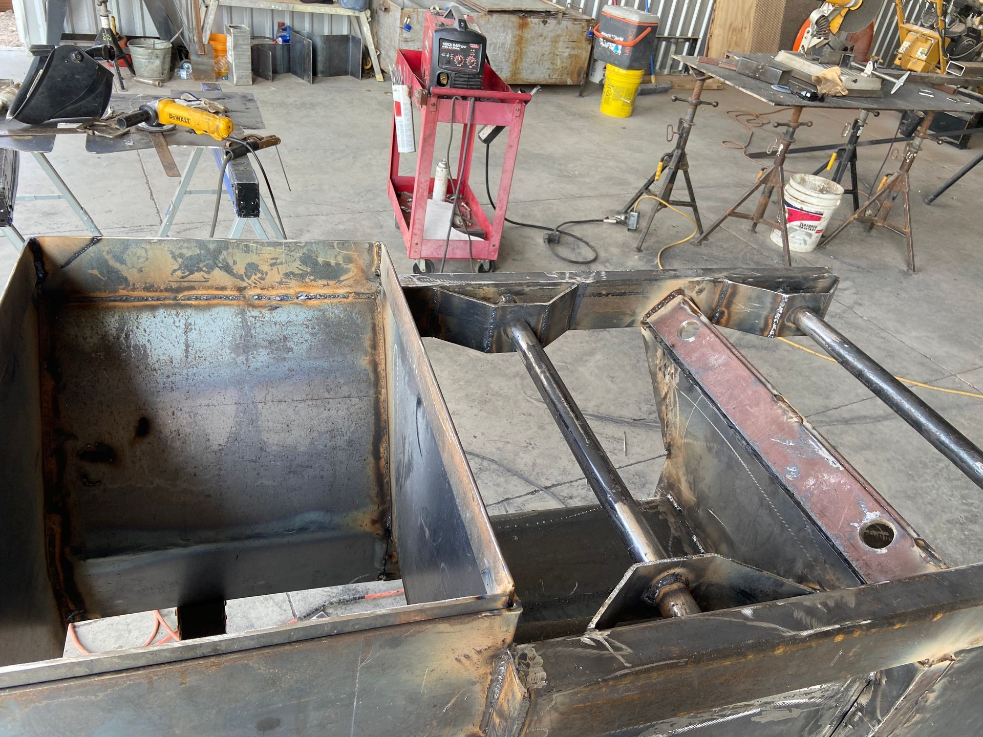 Welding workspace with metal structures in progress; red welding cart, tools, and equipment scattered around.