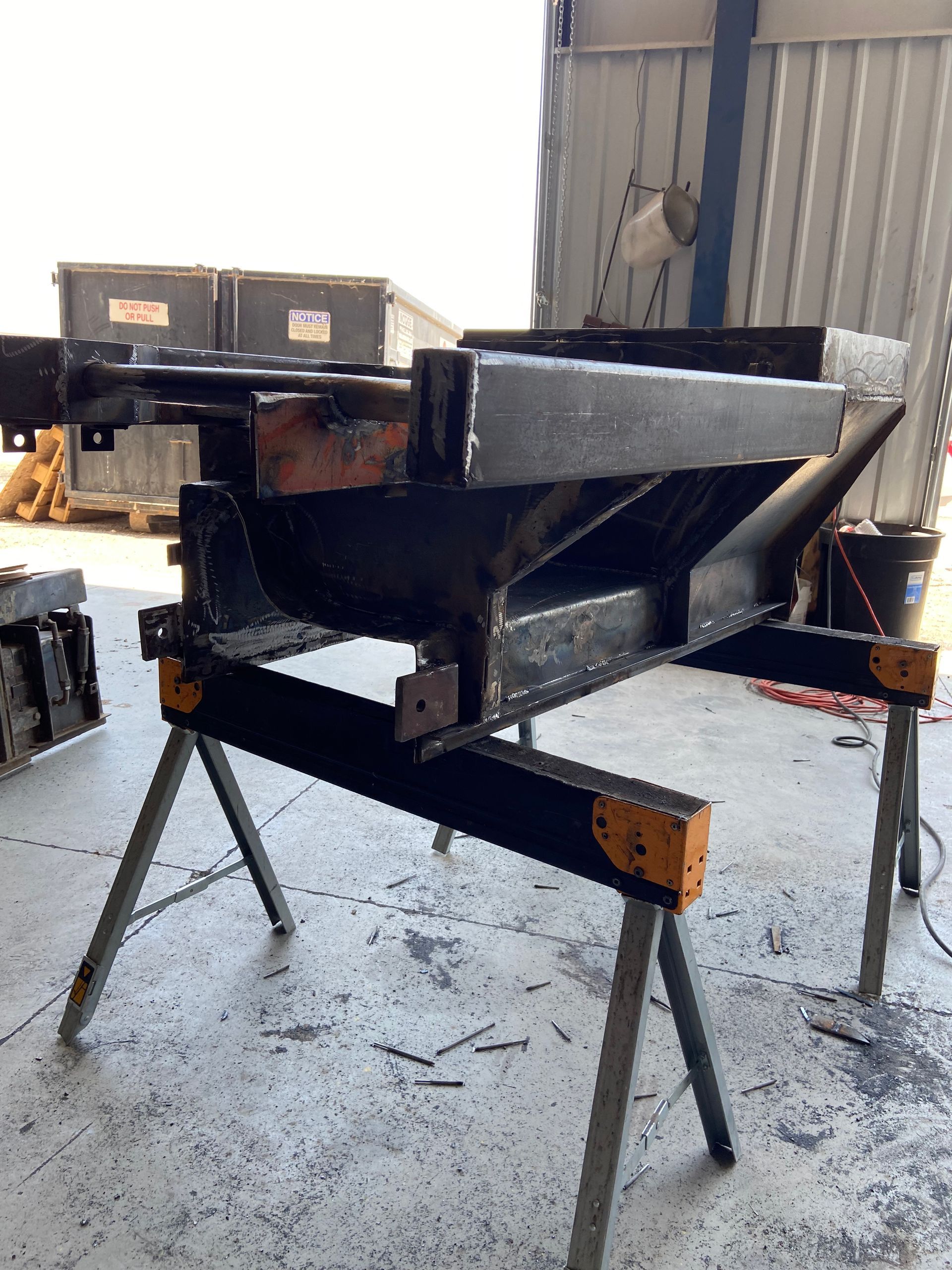 Black metal fabricated structure on orange and black sawhorses in a workshop setting.