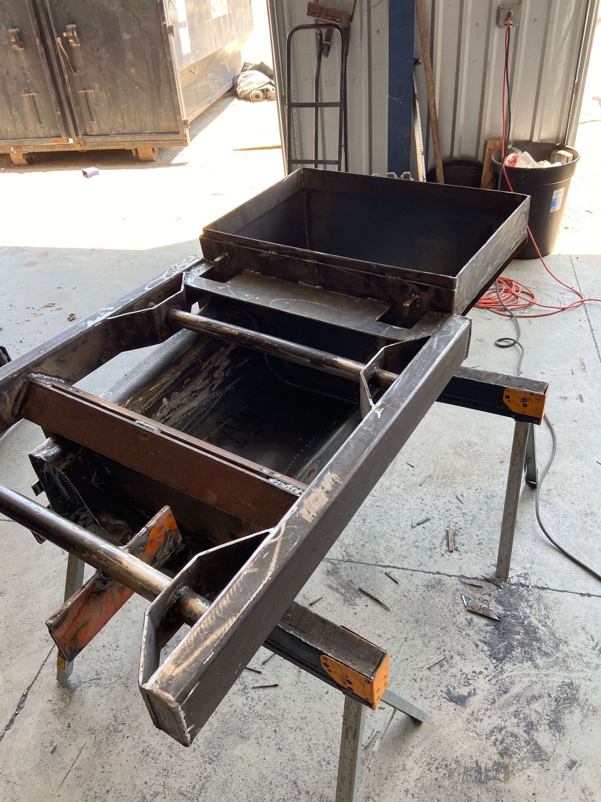 Metal fabricated sluice box, resting on two sawhorses in a workshop setting.