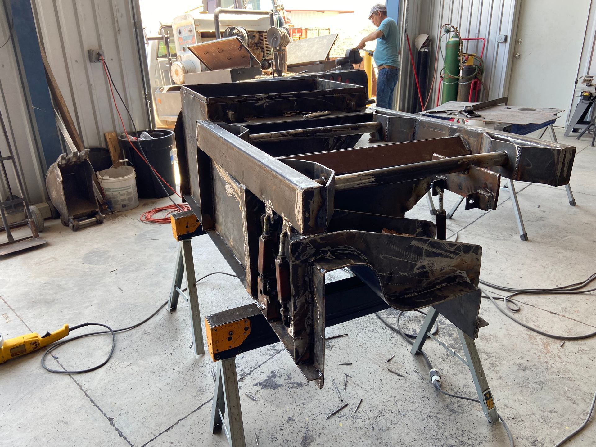 Welded metal frame on saw horses in a workshop; a person is welding in the background.