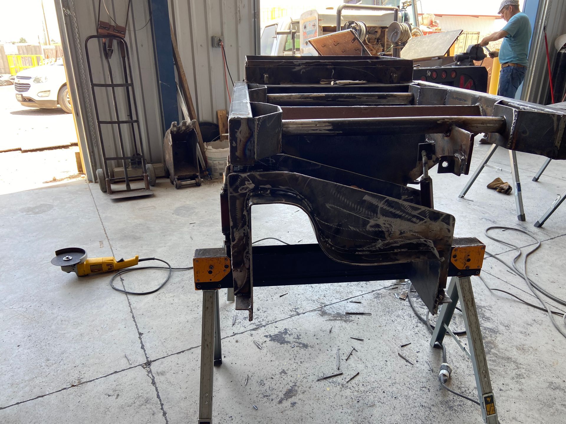 A metal frame on sawhorses in a workshop with a person in the background. A grinder is on the floor nearby.