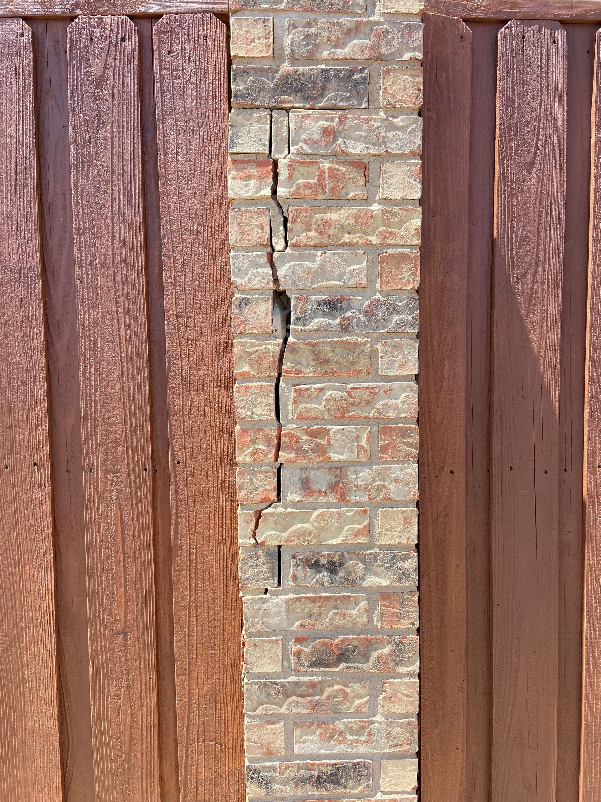 Brick pillar with large vertical crack, flanked by brown wooden fence panels.