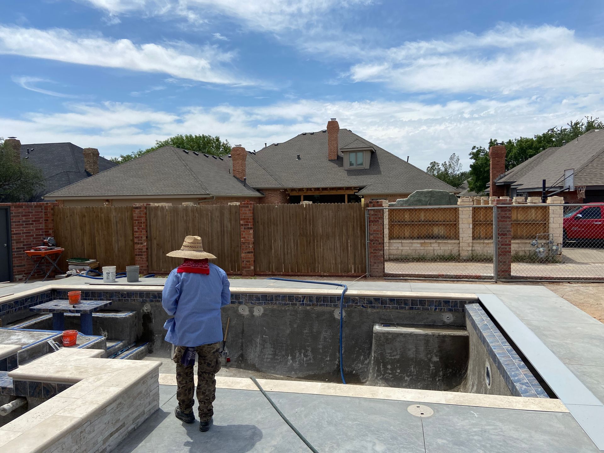 Person in a straw hat and blue shirt walks toward an empty pool, with houses and fence in background.