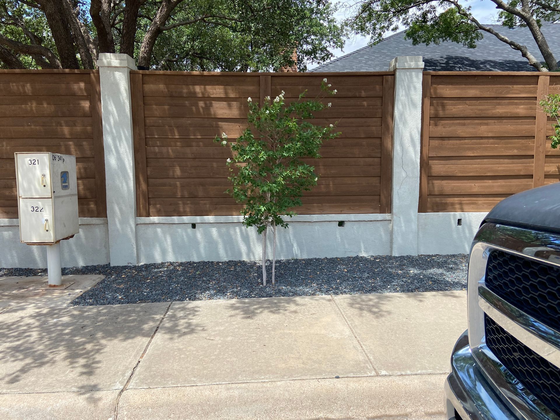 Brown wooden fence with white pillars, a young tree, and utility box.