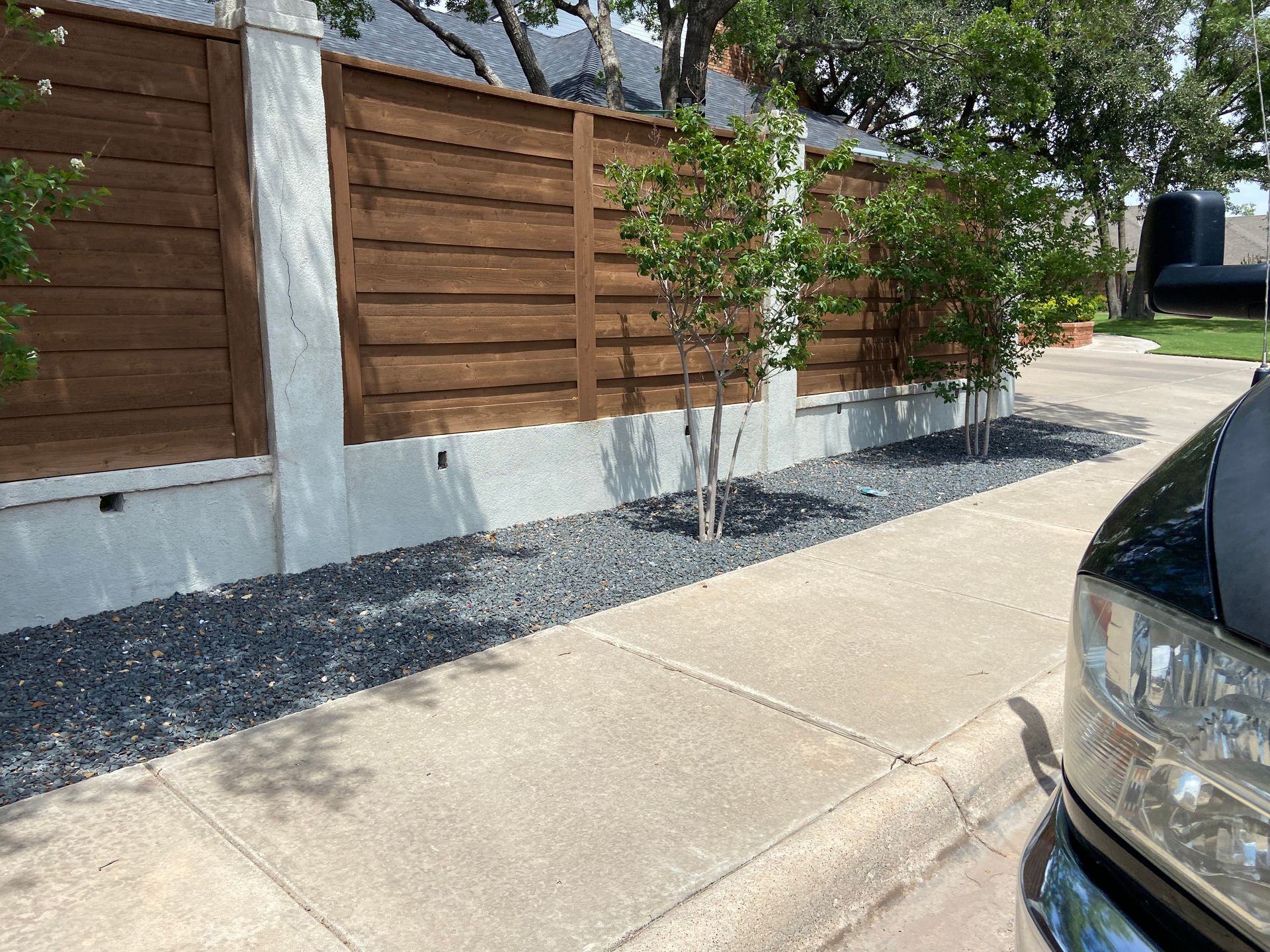 Sidewalk with gravel landscaping bed, trees, and wooden fence. Car parked on right.