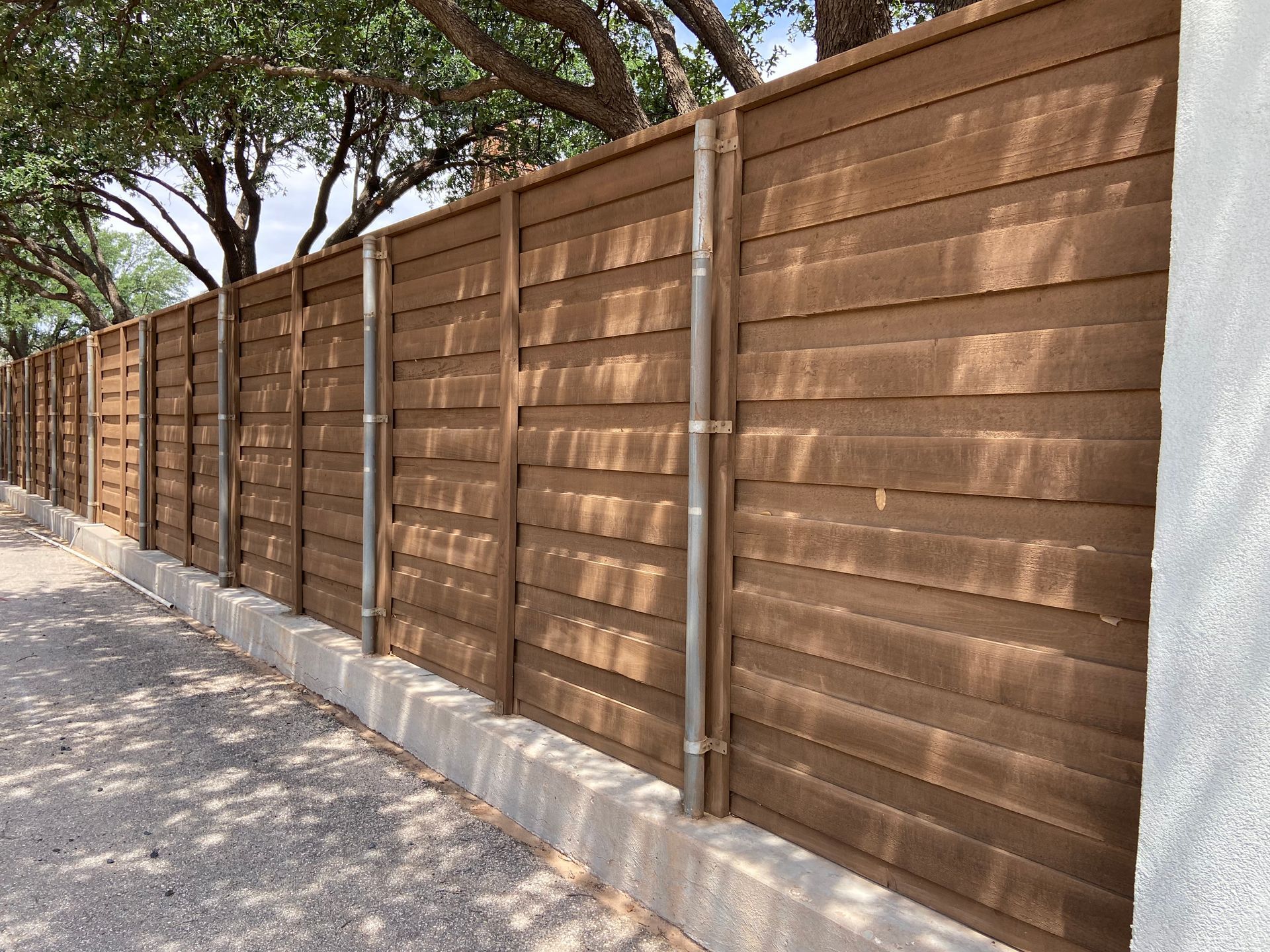 Brown horizontal plank fence with metal posts, next to a concrete curb and a gravel path.