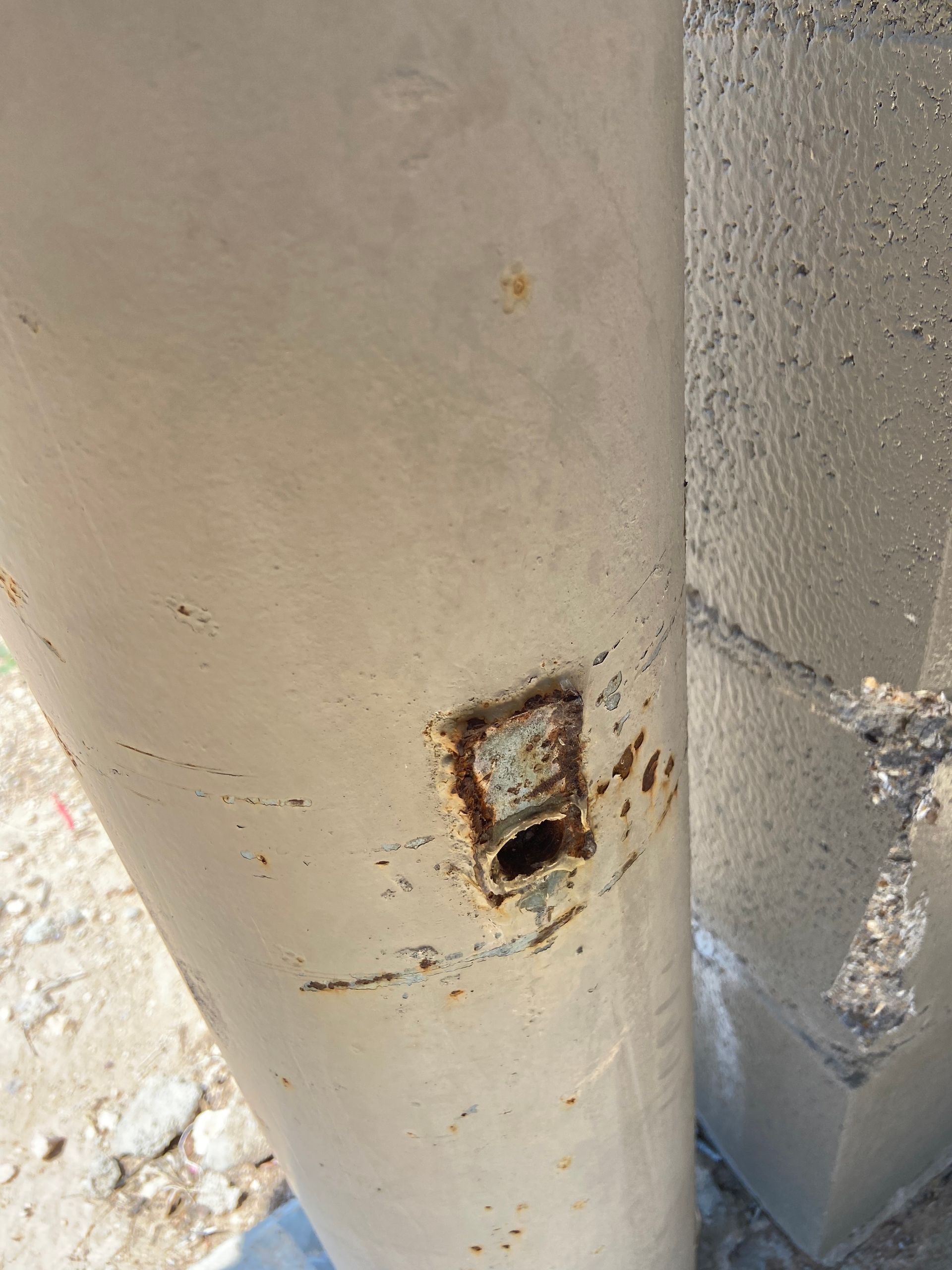 Rusting metal pole with a rectangular hole and discoloration, next to a concrete block.