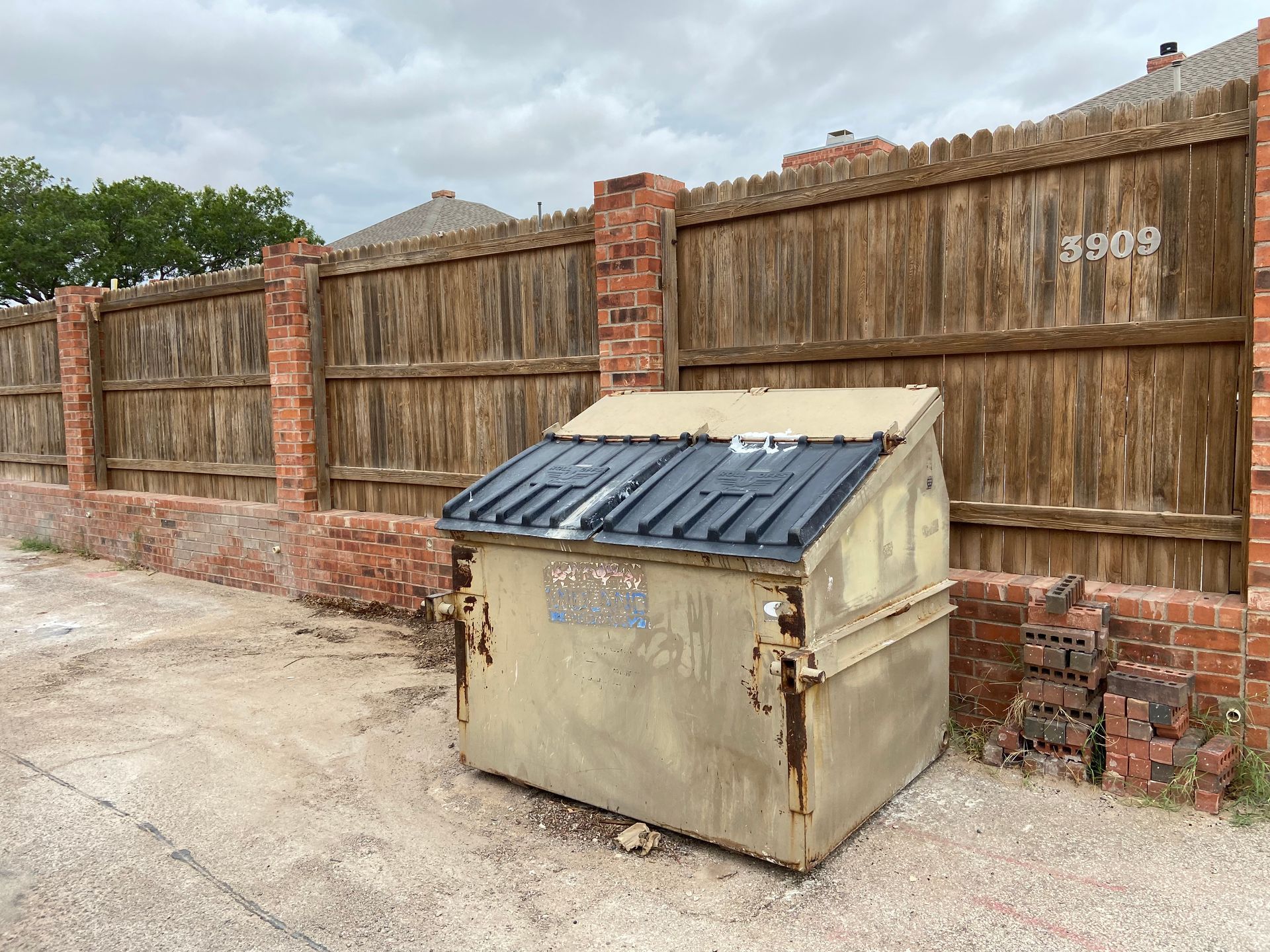 A weathered dumpster in front of a wooden fence with brick columns. Cloudy sky overhead.