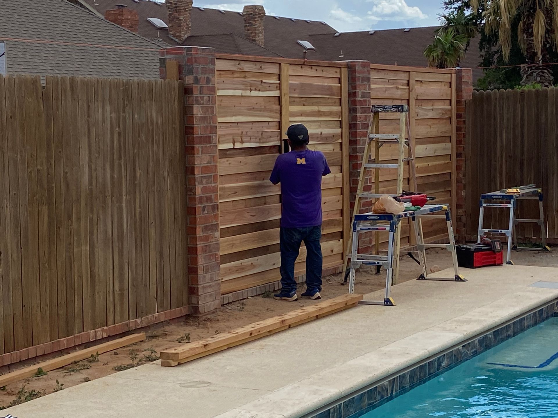 Man building a wooden fence next to a brick pillar and swimming pool.