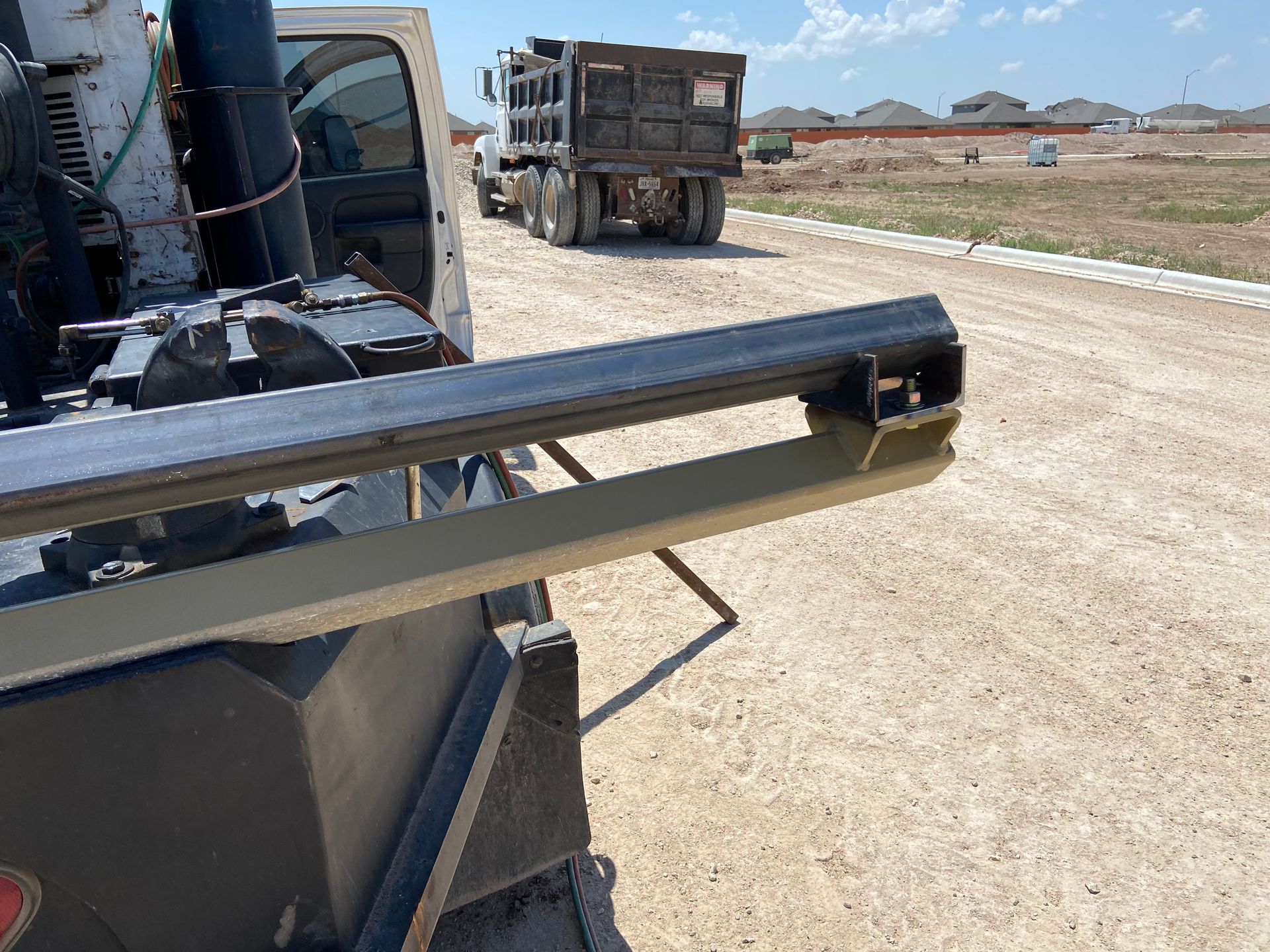 Close-up of a metal beam protruding from a truck bed, with a dump truck and construction site in the background.
