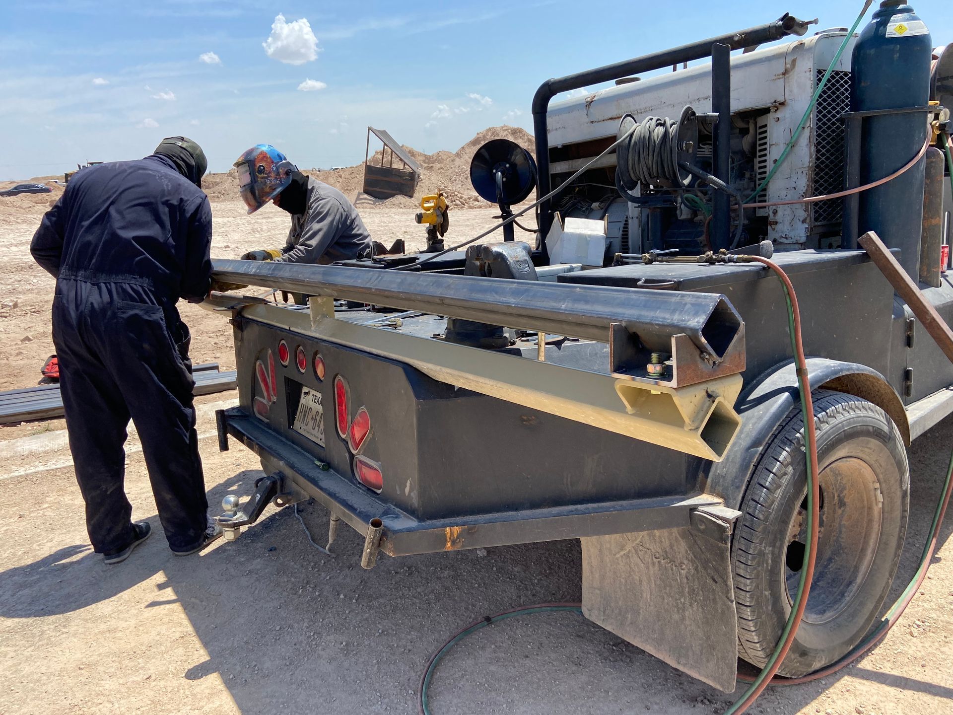 Two workers welding on a trailer bed in a desert environment under a blue sky.