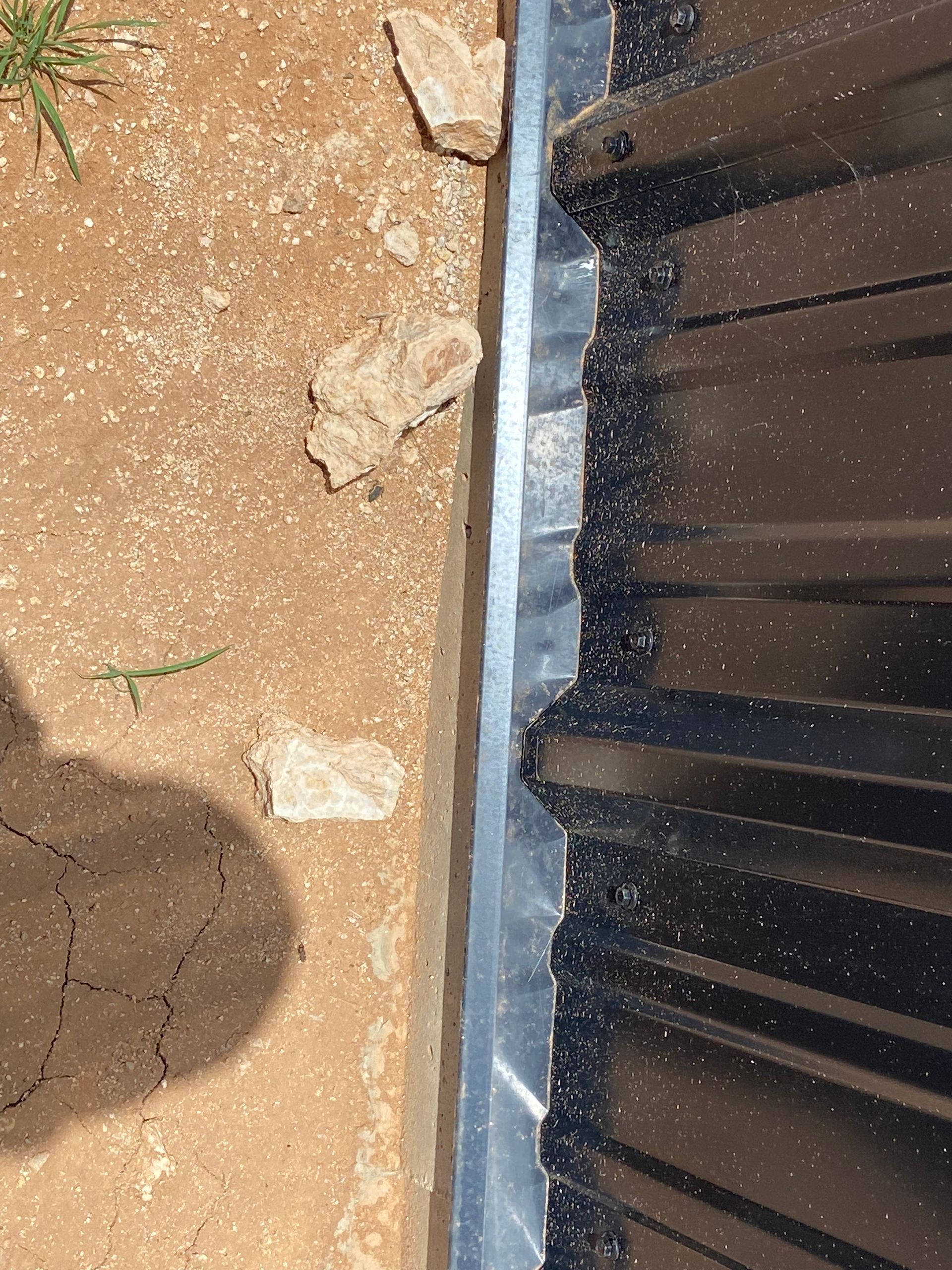 Metal roofing edge next to a dirt area, with rocks and some vegetation visible.