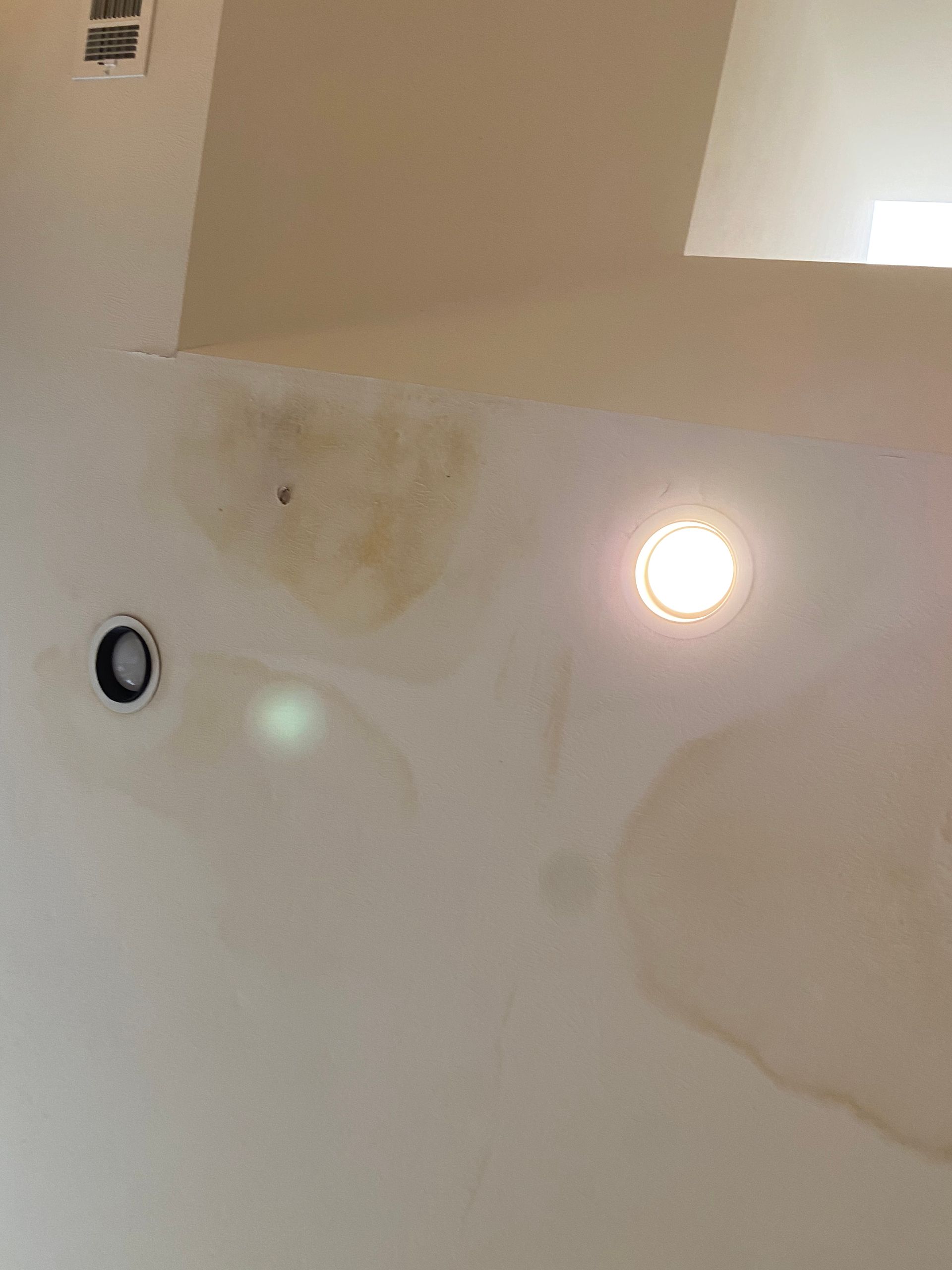 Water damage stain on a light-colored ceiling with two recessed lights and an air vent.