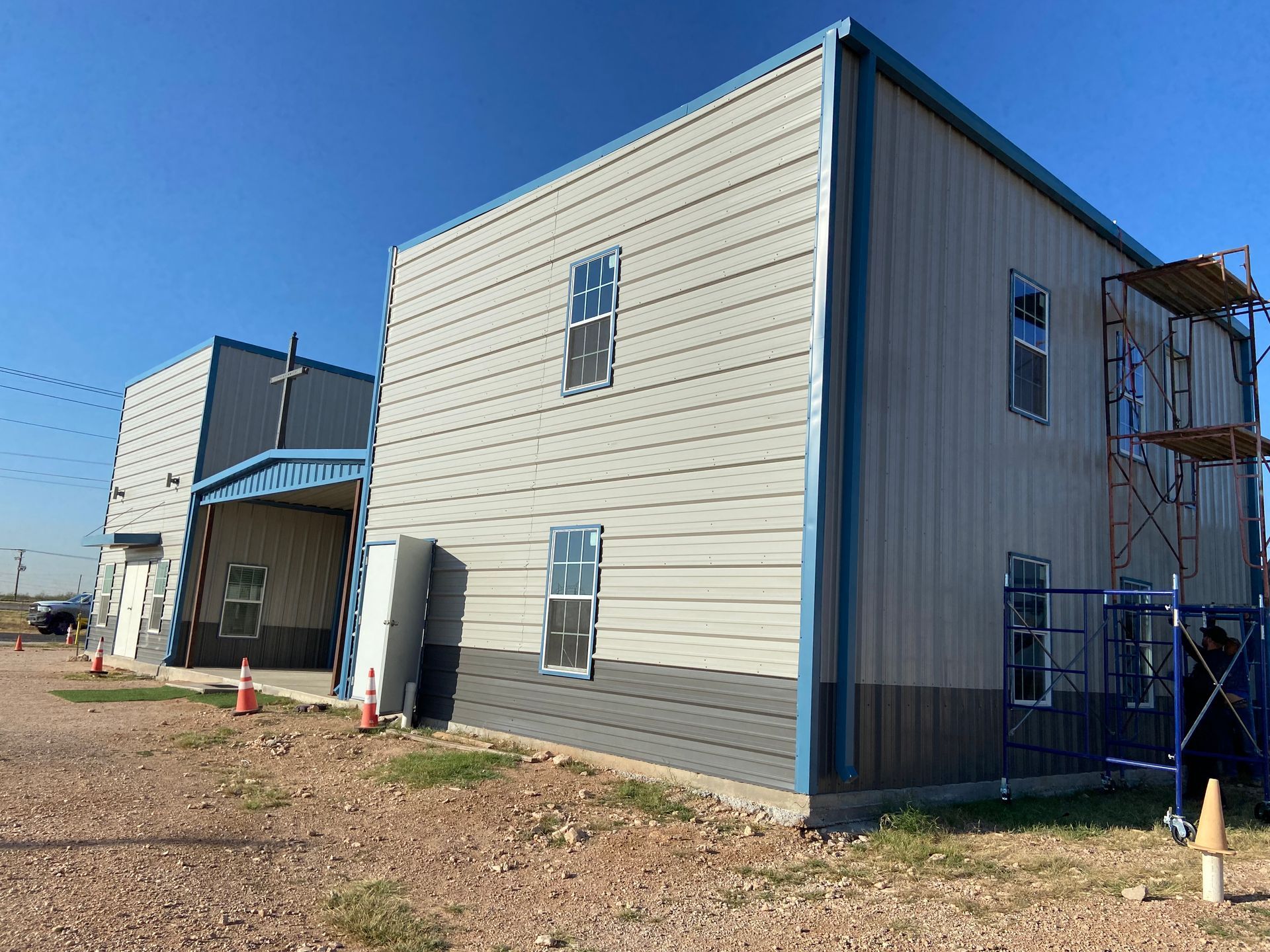 Two-story metal building under construction, light gray siding, blue trim, scaffolding, clear sky.