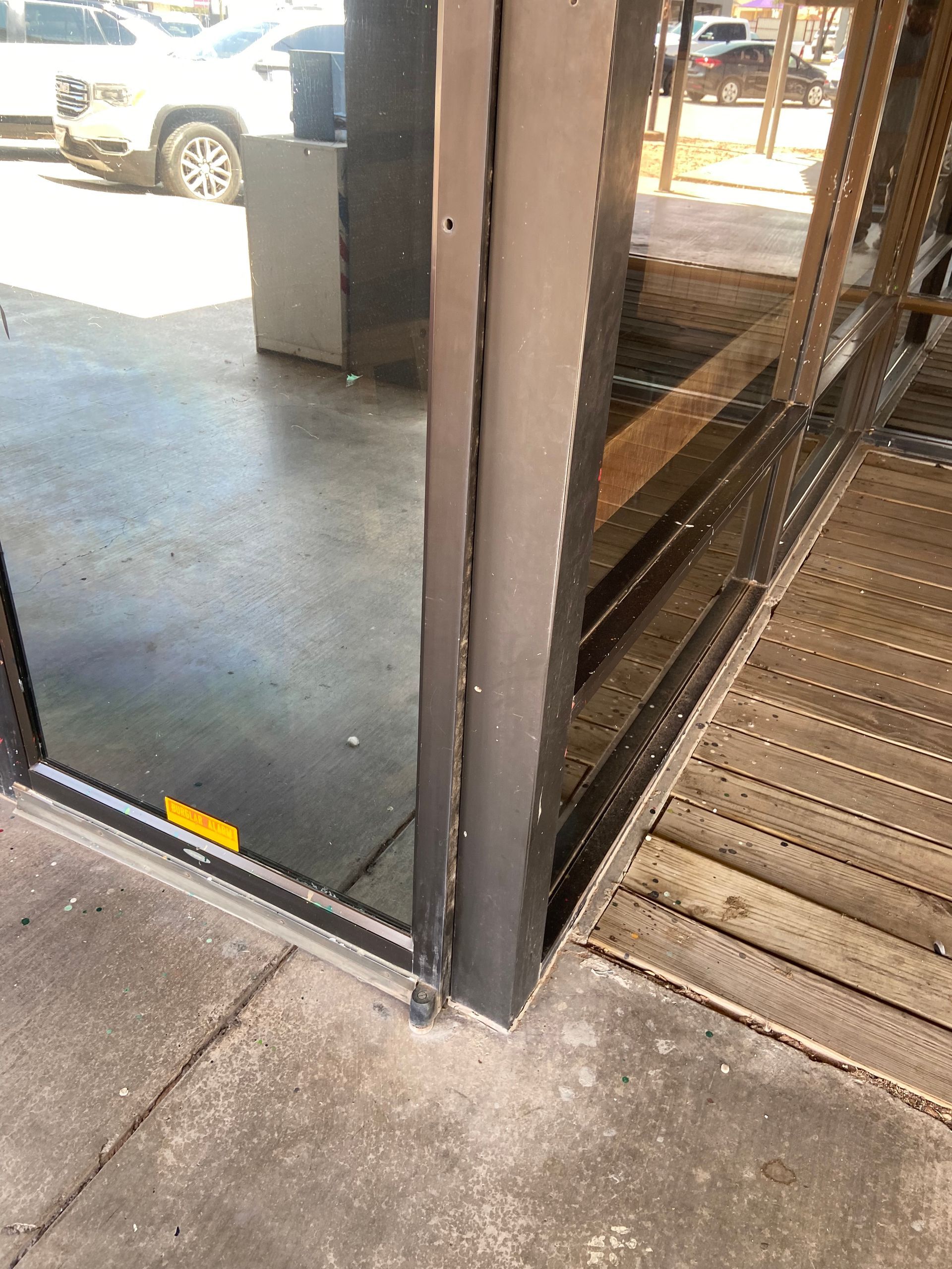 Corner of a building with a glass window and a wooden deck; a sidewalk is in front.