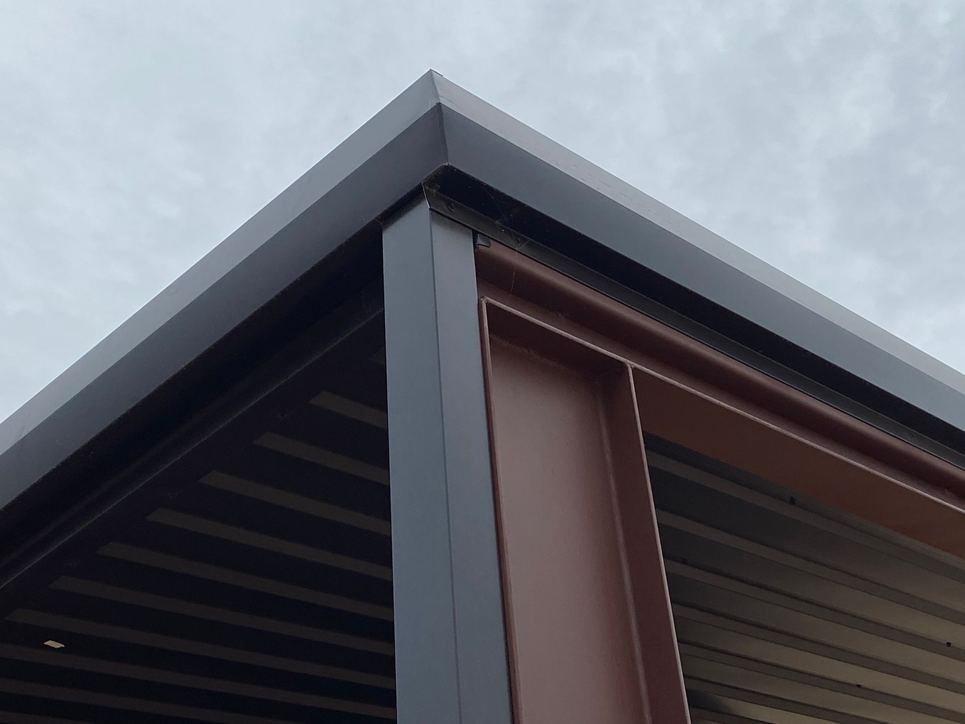 Corner of a modern, dark-colored building with a brown support beam against a cloudy sky.