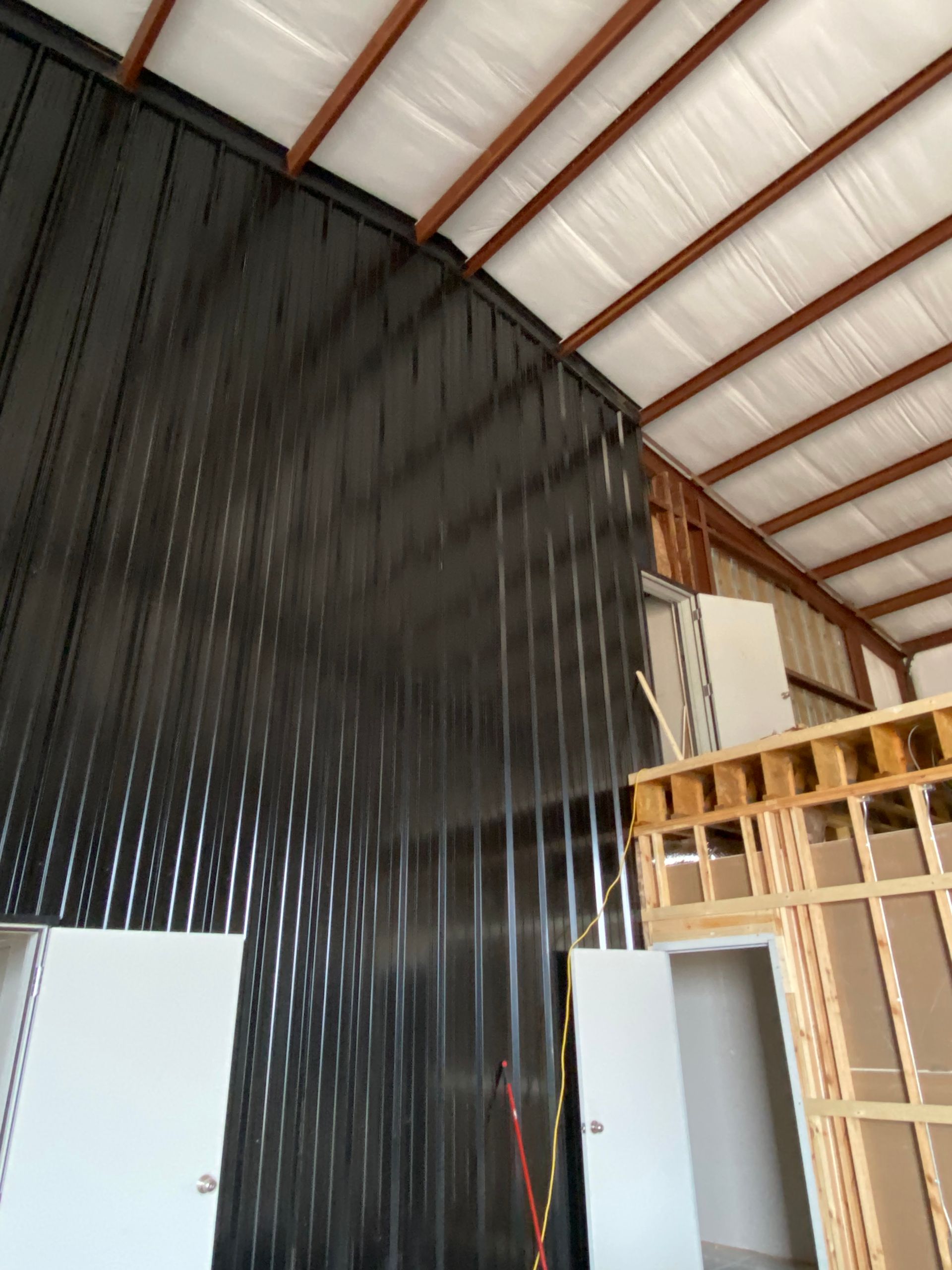Interior view of a metal building with black walls, open rafters, and wood framing. White doors are visible.