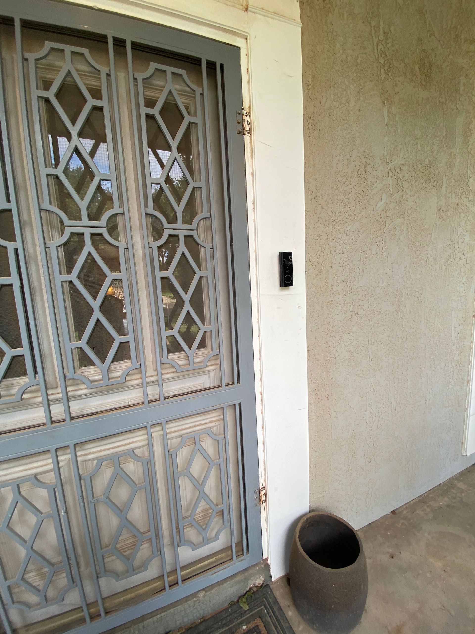Exterior view of a door with decorative gray security bars, a black doorbell, and a dark brown planter.