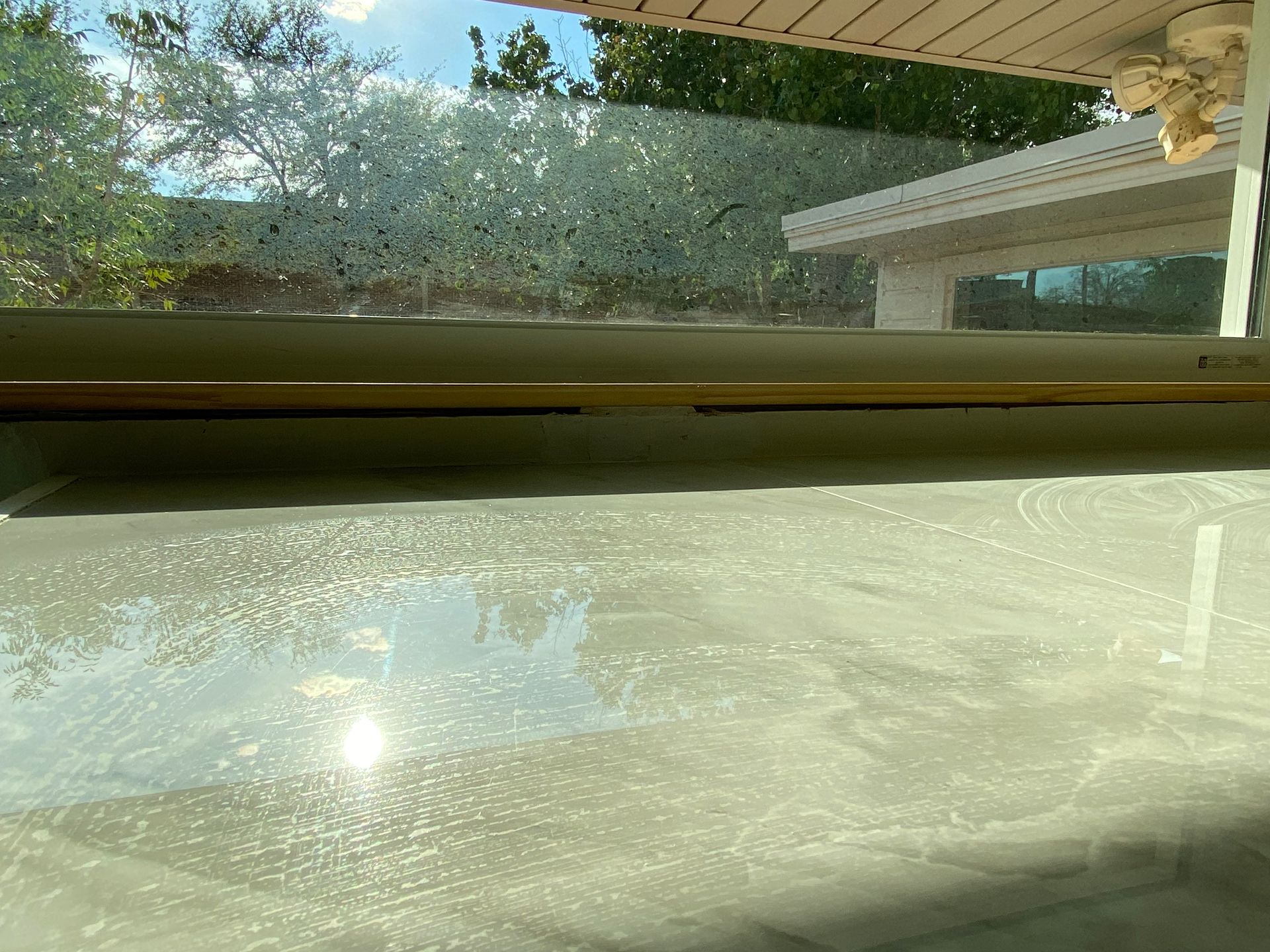 Window with condensation, reflecting the sun and a partial view of a covered patio and trees.
