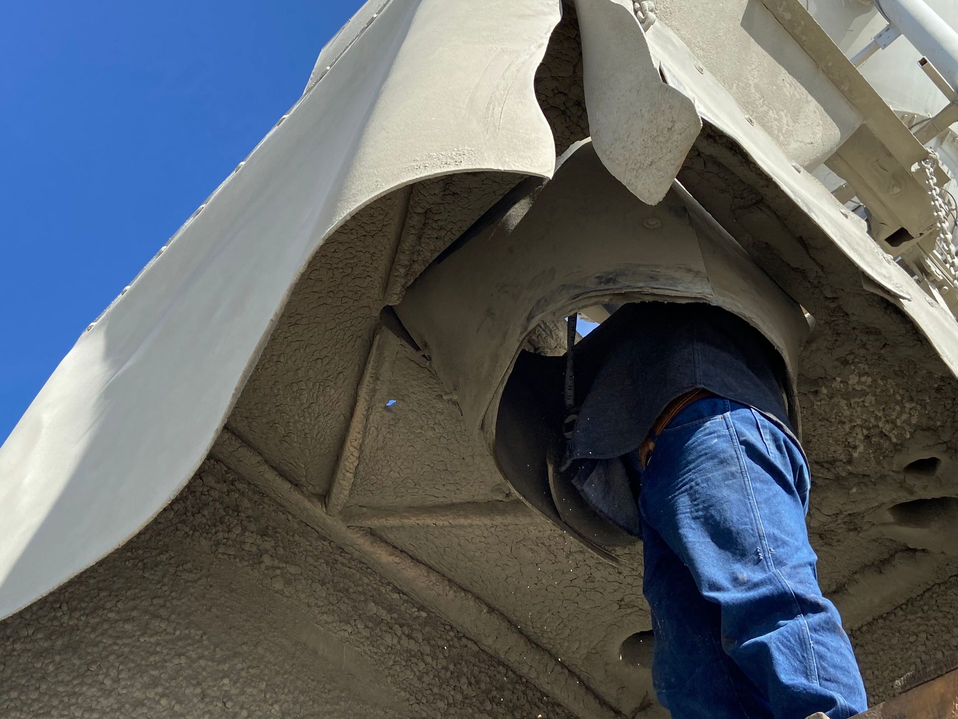 Person in blue jeans working under a concrete mixer drum, sunlight, sky.