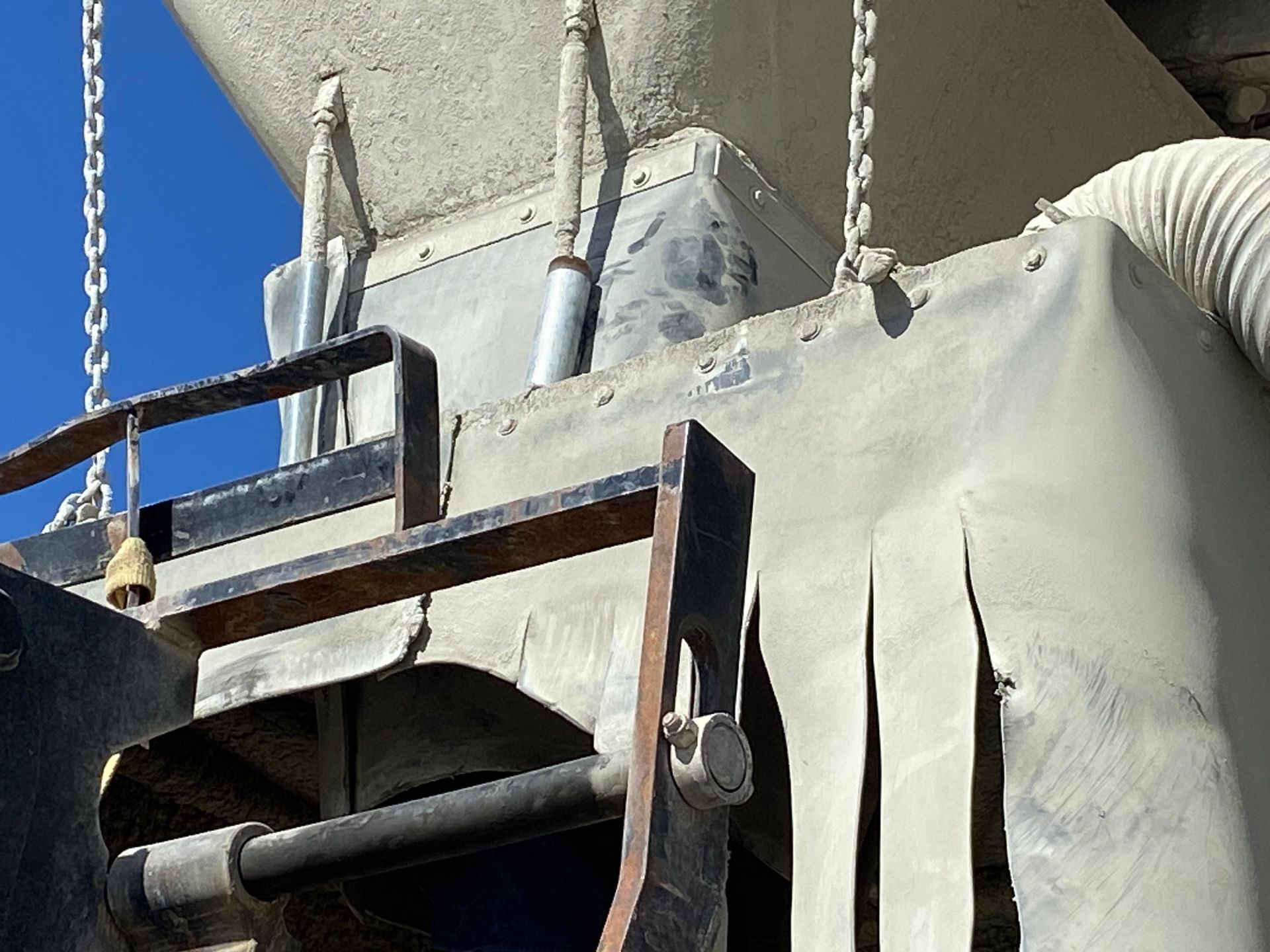 industrial machinery with torn, light-colored covering, metal chains, and a white, ribbed hose against a blue sky.
