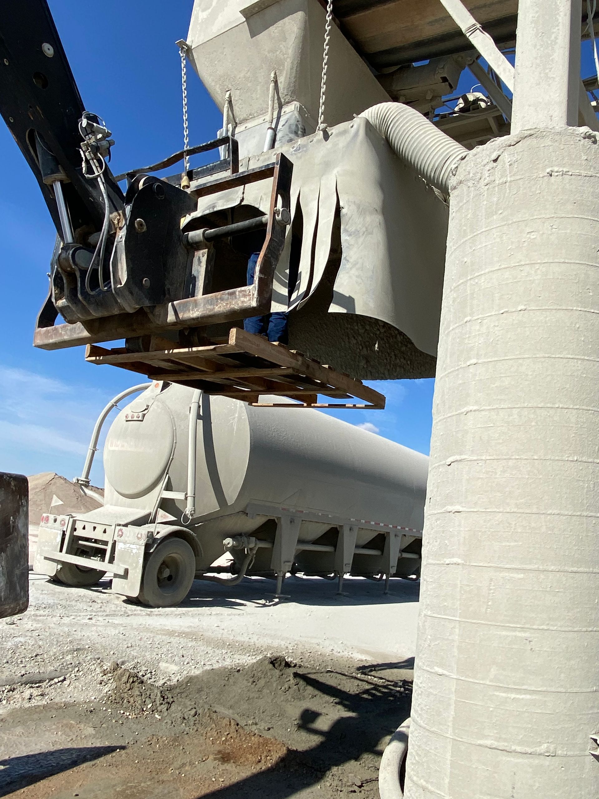 Excavator dumping material into a concrete silo, with a tanker truck in the background. Bright sunny day.