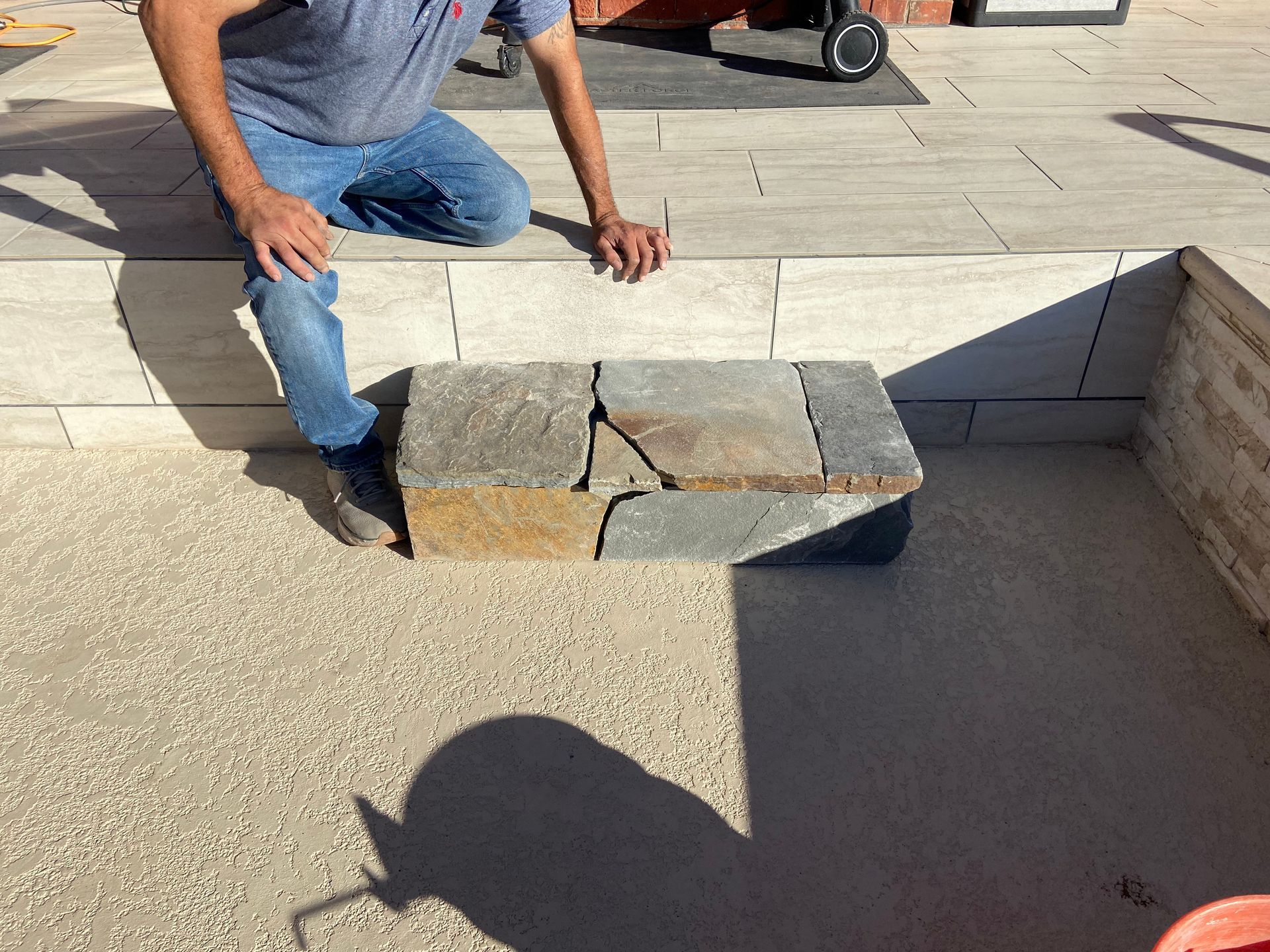Person kneeling by a stone-covered seating area on a paved patio. Sunny outdoor setting.