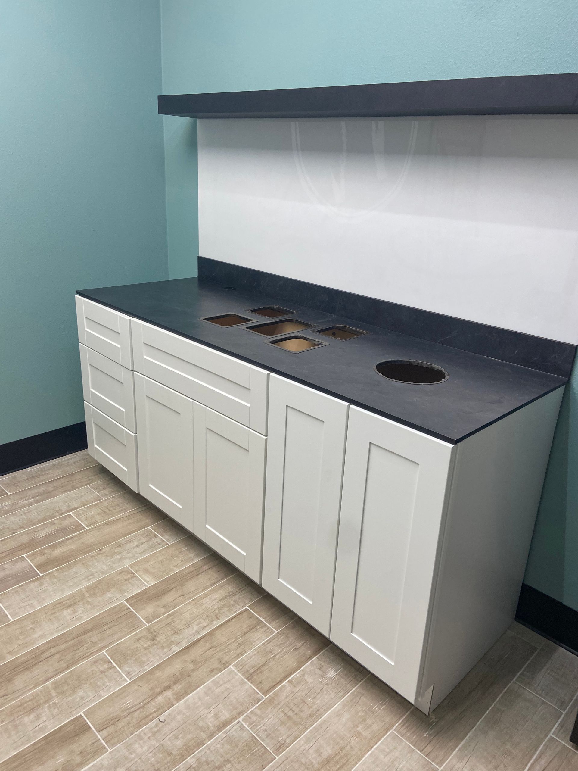 White cabinets with a dark countertop and holes, against a teal wall. Wooden floor.