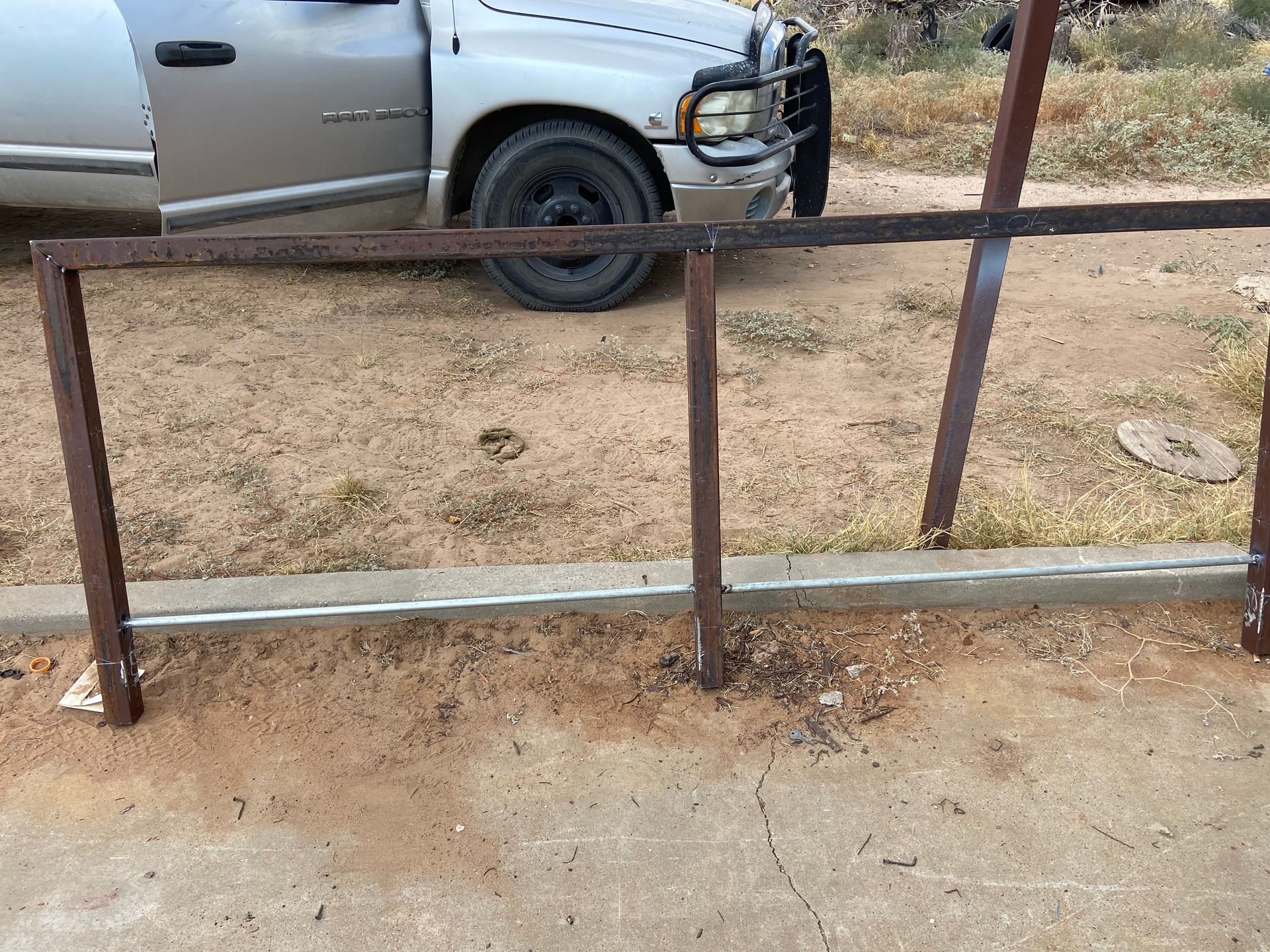 Metal frames of a fence welded to vertical supports, with a truck in the background on a dirt surface.
