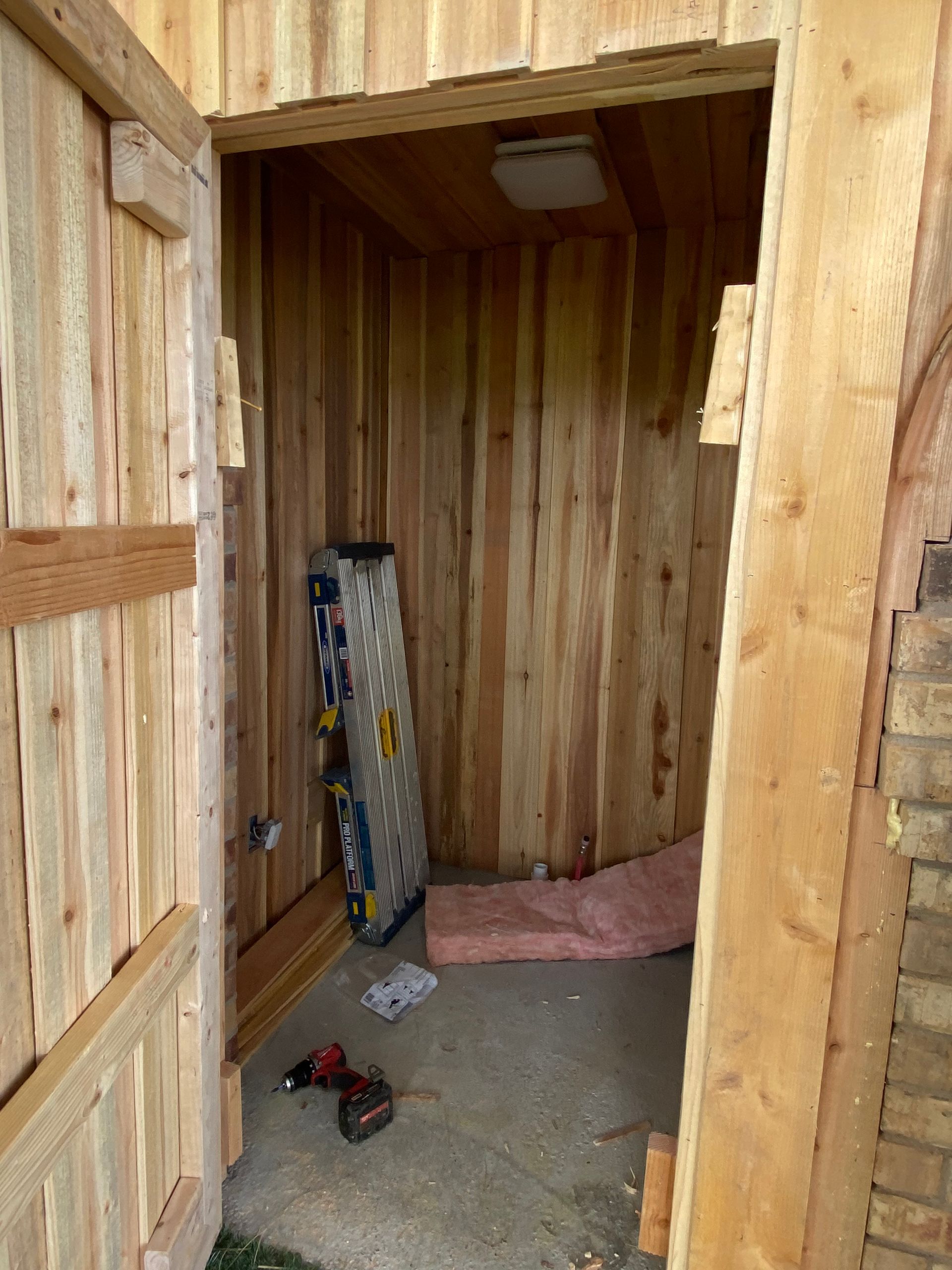 Wooden shed interior with insulation, tools, and a partially open door. Cedar plank walls.