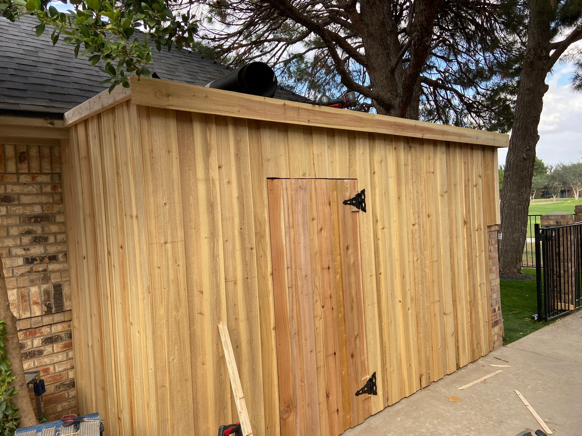 Wooden shed with a door and a flat roof attached to a brick wall next to a sidewalk and a tree.