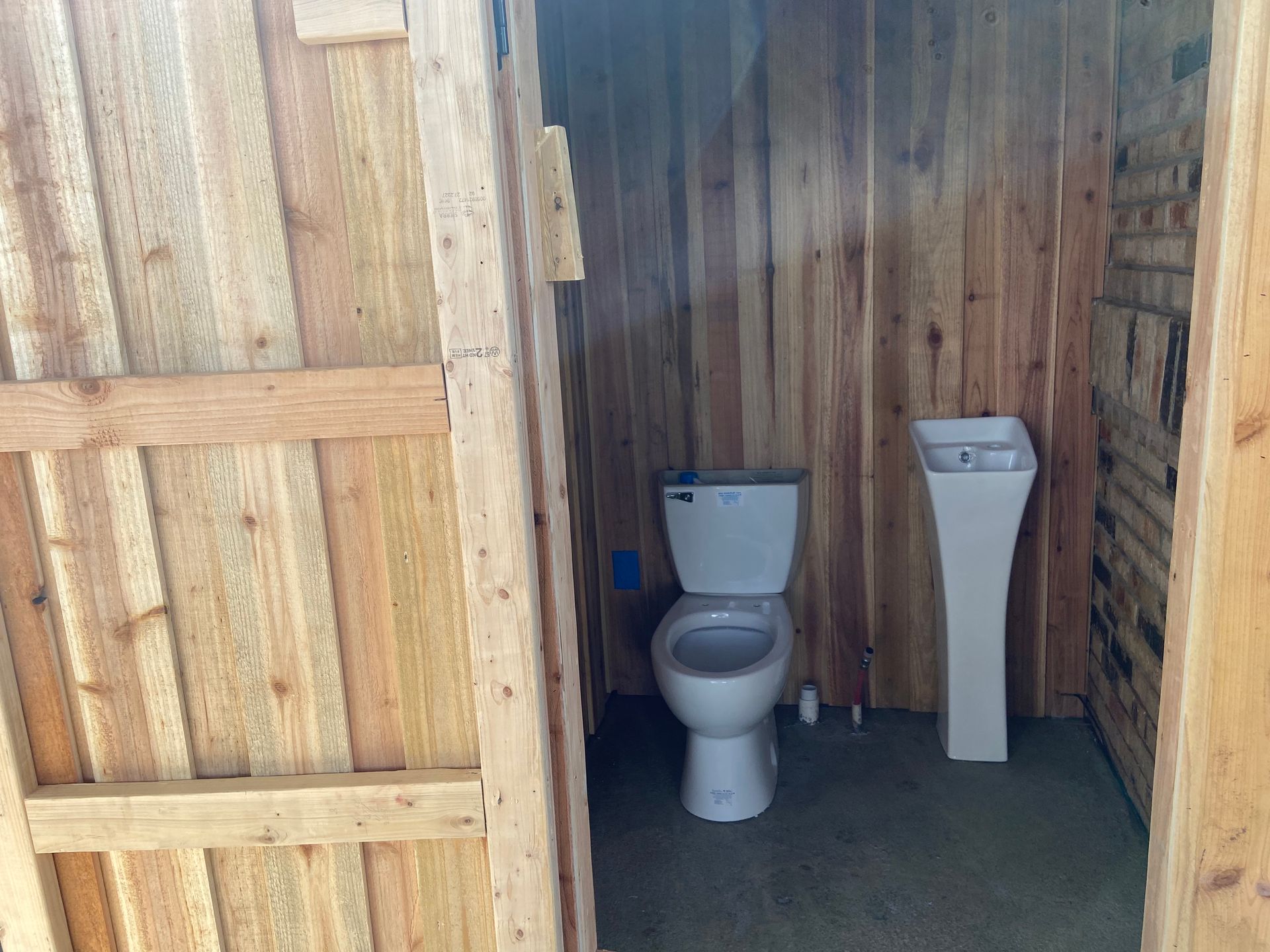 Interior of a wooden outhouse; toilet and pedestal sink are visible.