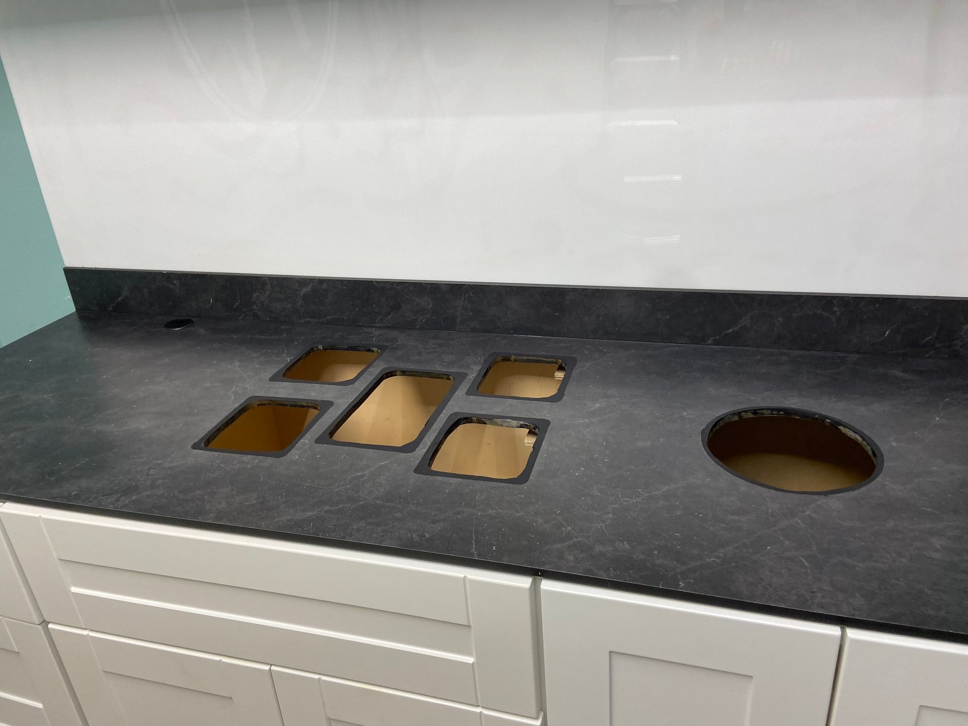 Dark countertop with cutouts for sink and fixtures on white cabinet, white tile backsplash.