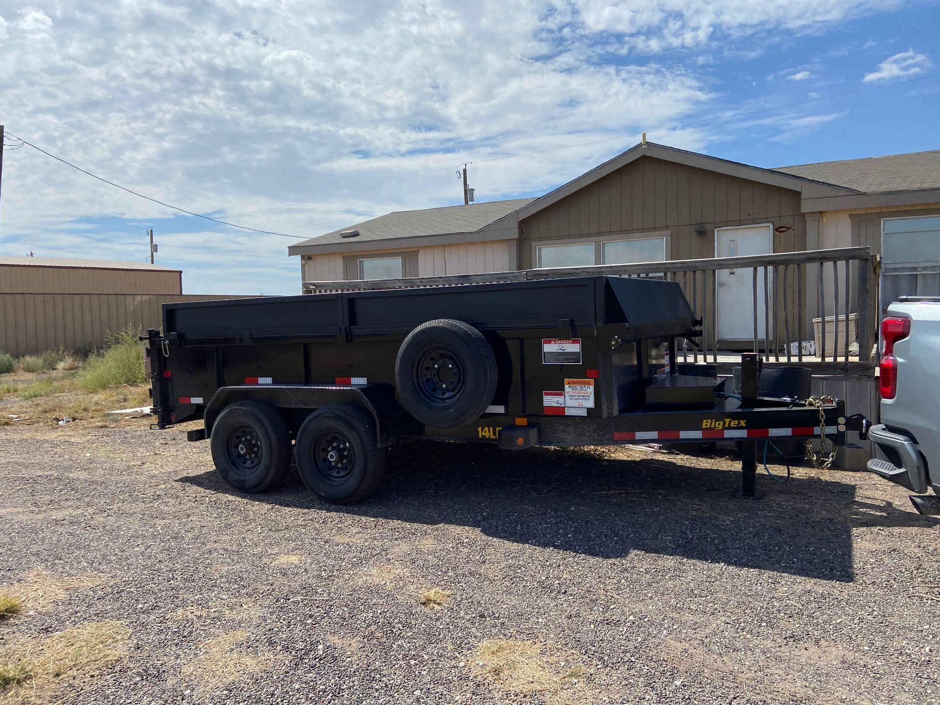 Black dump trailer parked on gravel in front of a house on a sunny day.