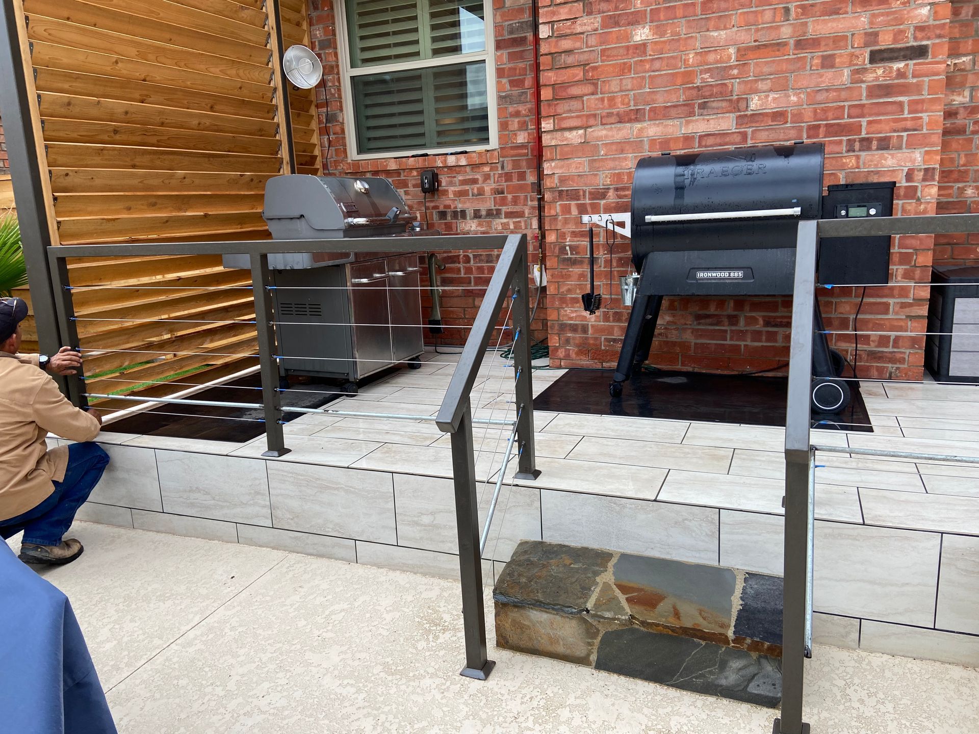 Man installing railing on a patio with two grills. Brick wall, wooden slats, and stone steps are visible.