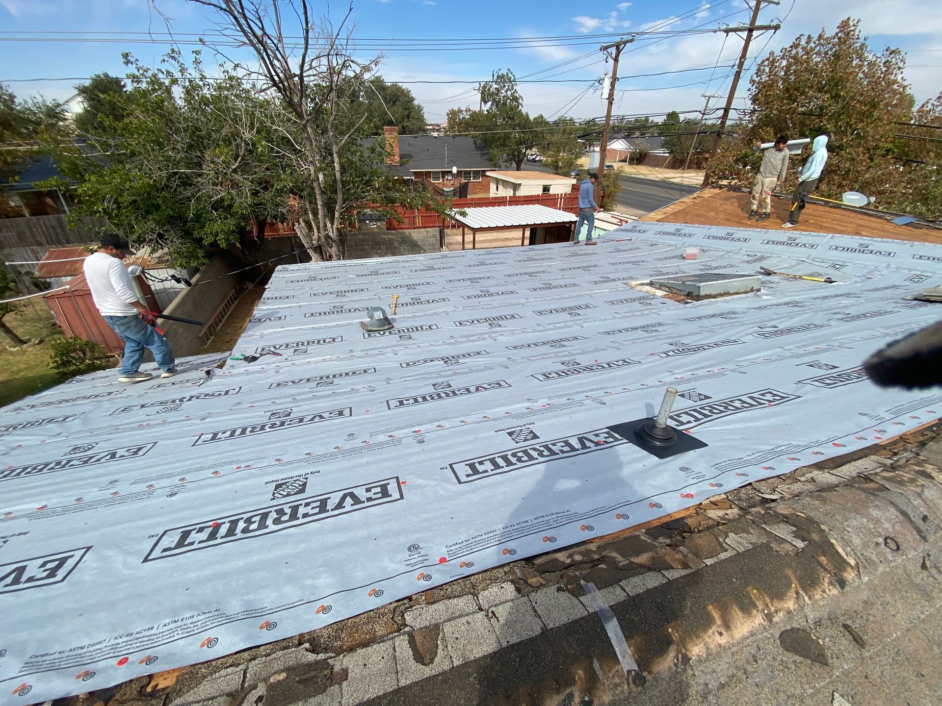 Workers on a rooftop, applying roofing material. Blue sky, houses in the background.