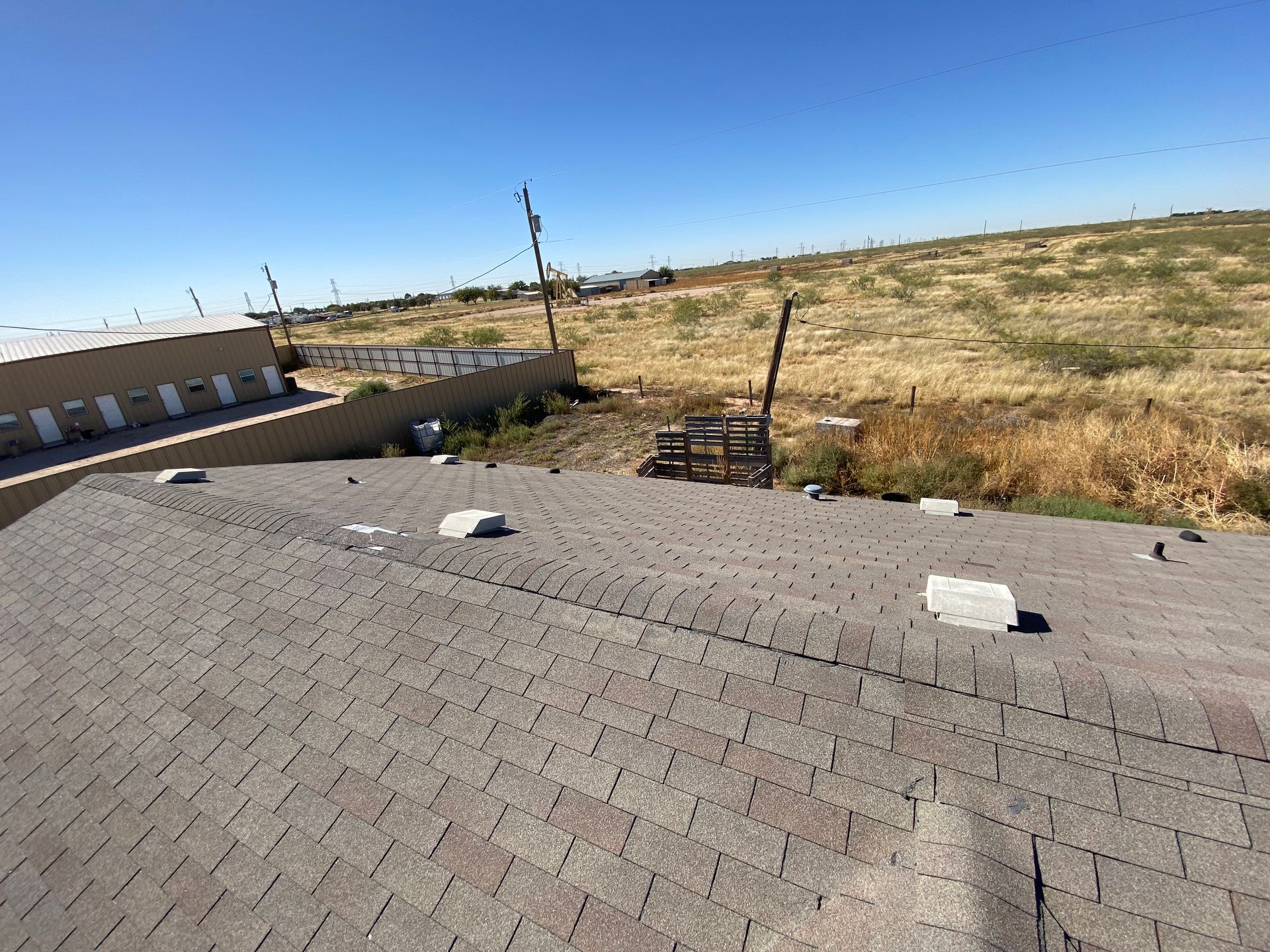 View from a roof, showing shingles, buildings, field, fence, power lines under a blue sky.