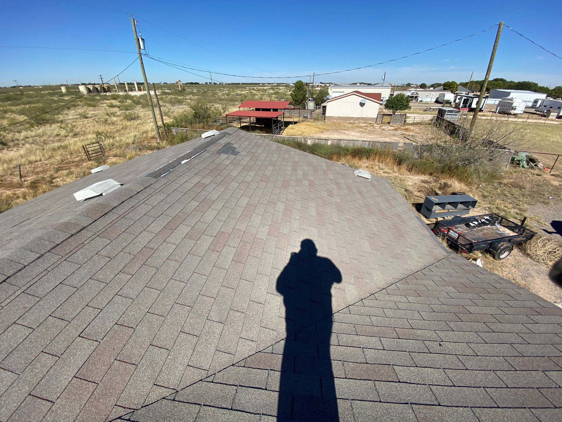 Shadow of person on shingled roof overlooking rural buildings and landscape under a bright sky.