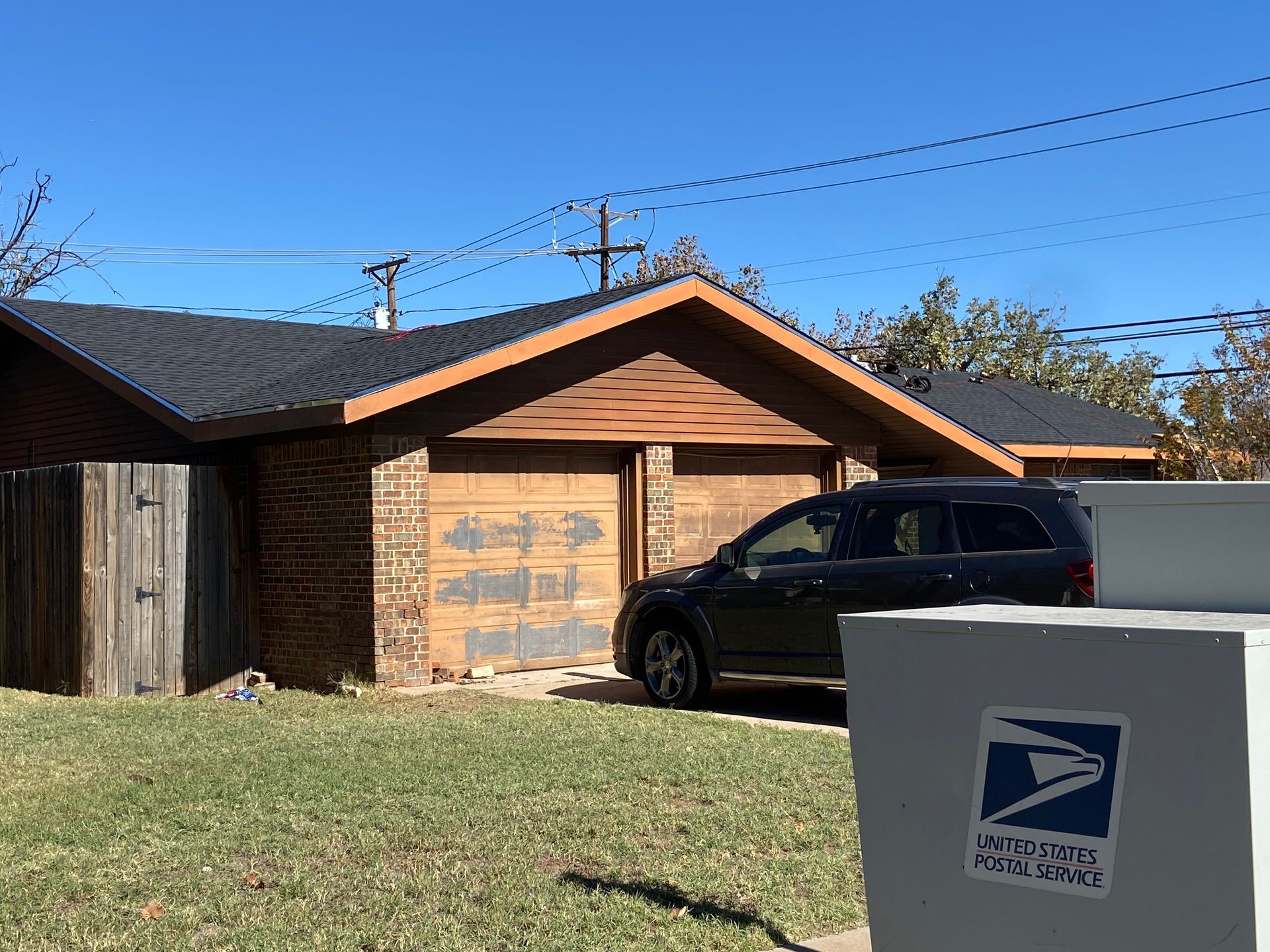 Black car parked in front of a brick garage with brown doors, mailbox in the foreground, sunny day.