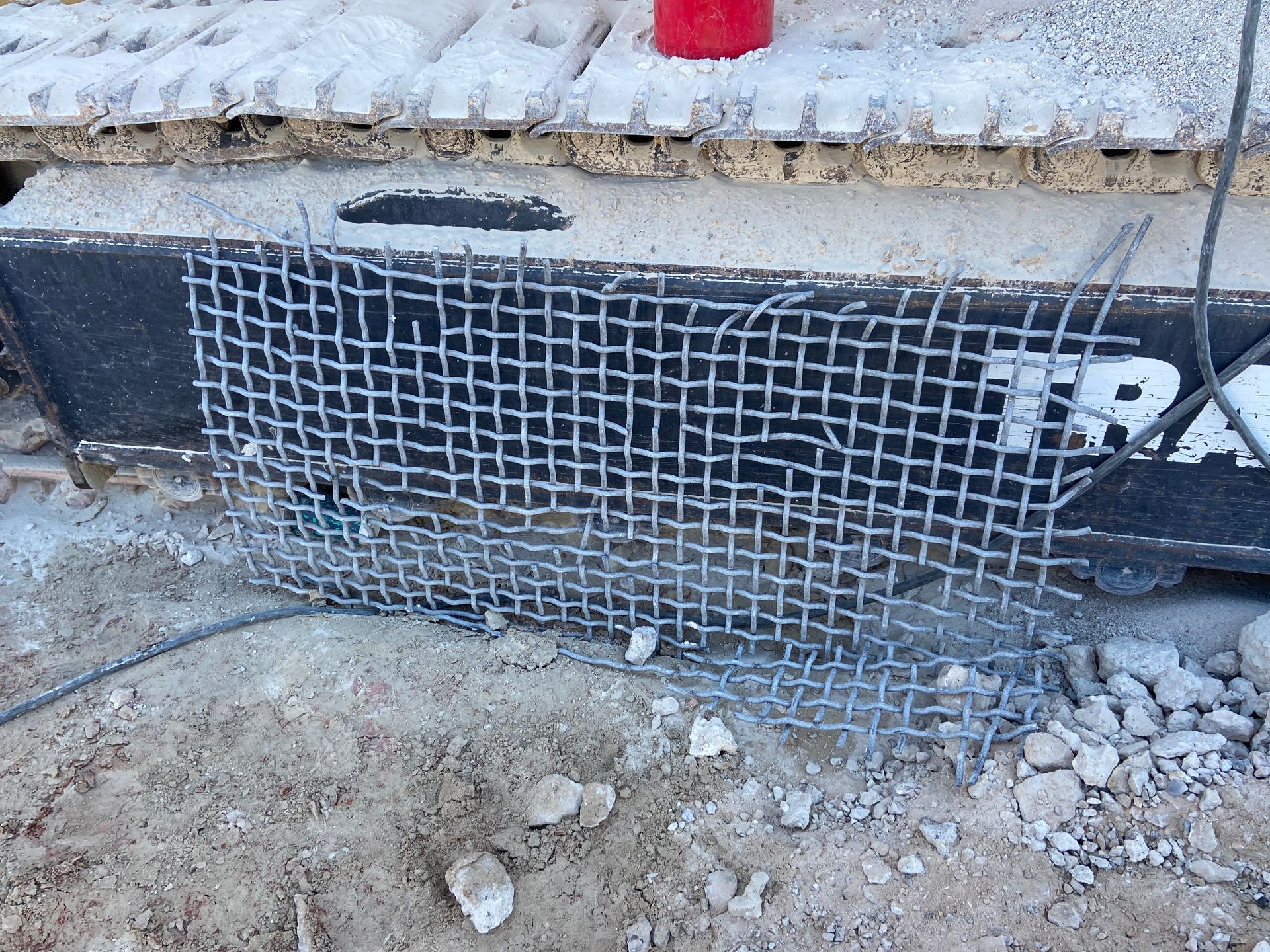 Wire mesh placed in front of machinery tracks on dirt, with debris around.