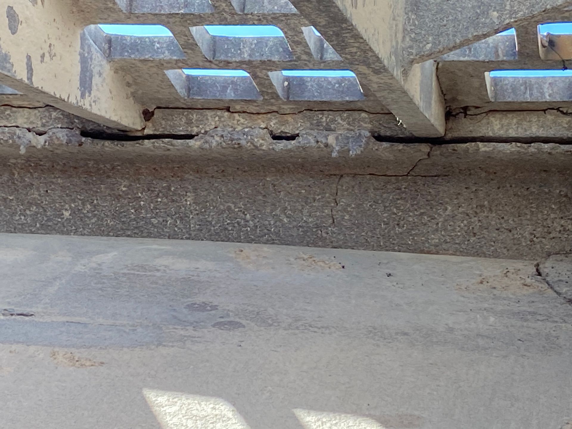 Close-up of a concrete structure with metal grating overhead. Cracks are visible in the concrete.