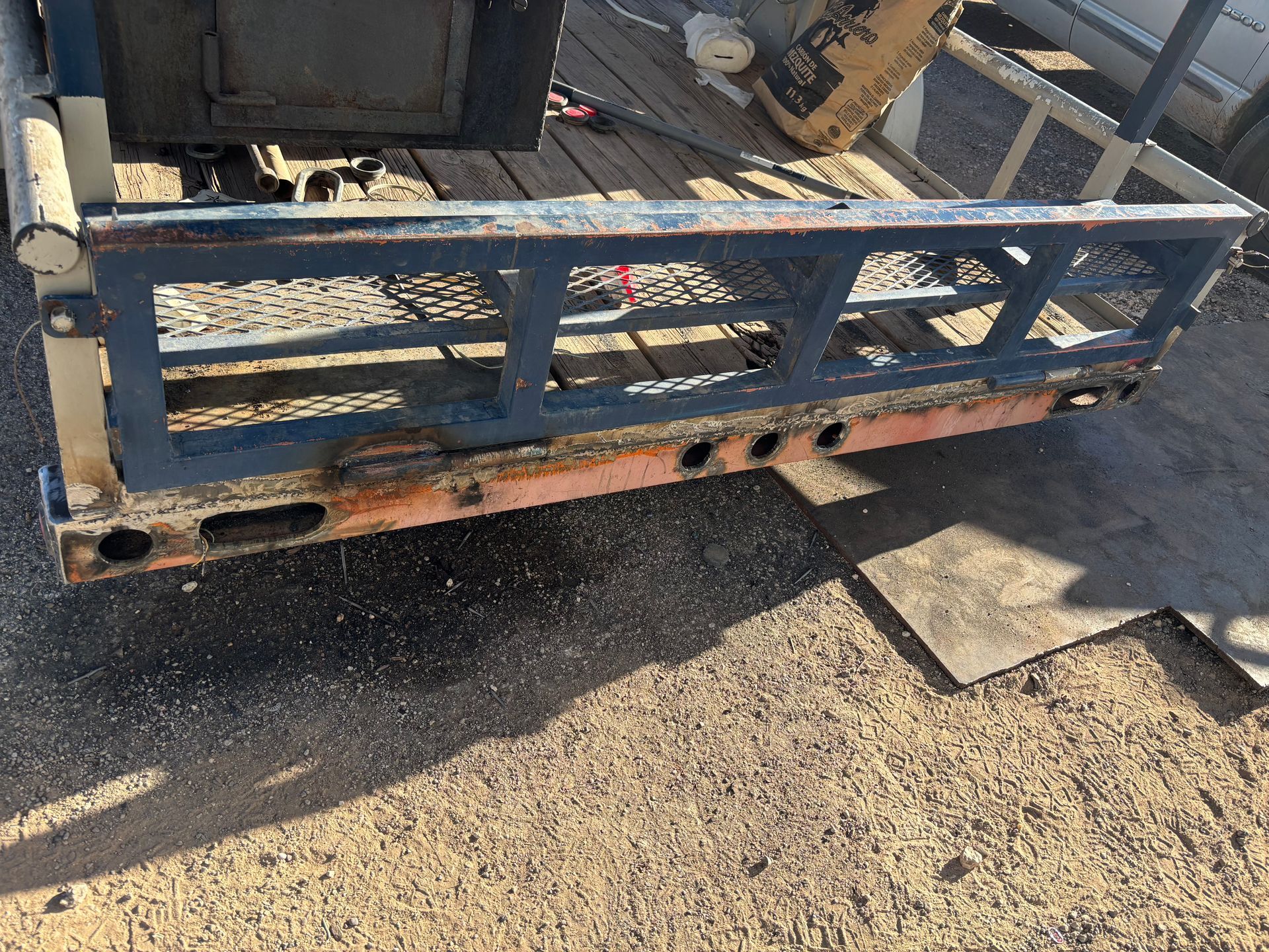 A blue and rusty metal utility trailer with a wooden bed sits on gravel in daylight.