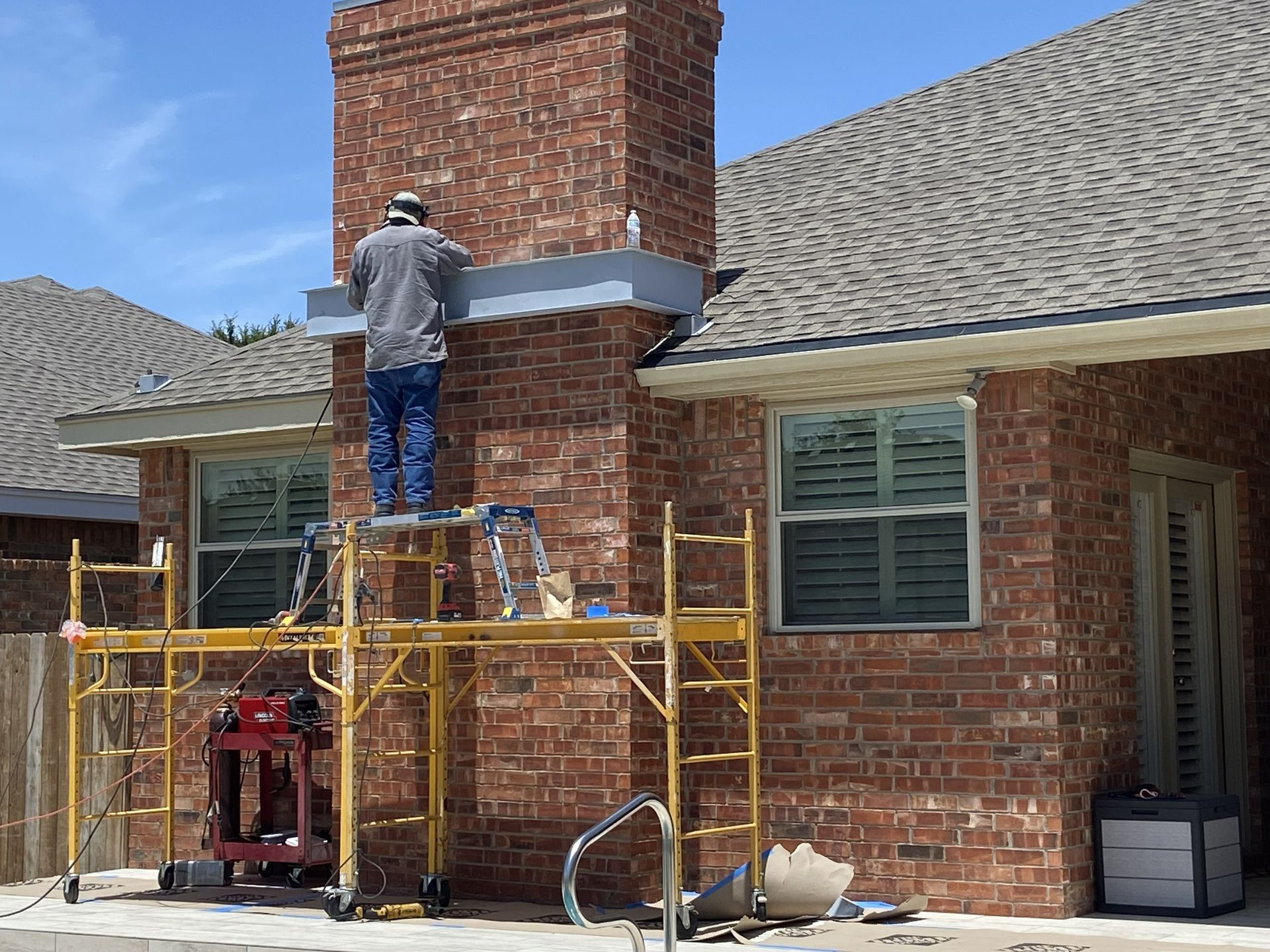 Person on scaffolding repairing a brick chimney next to a house with a pool.