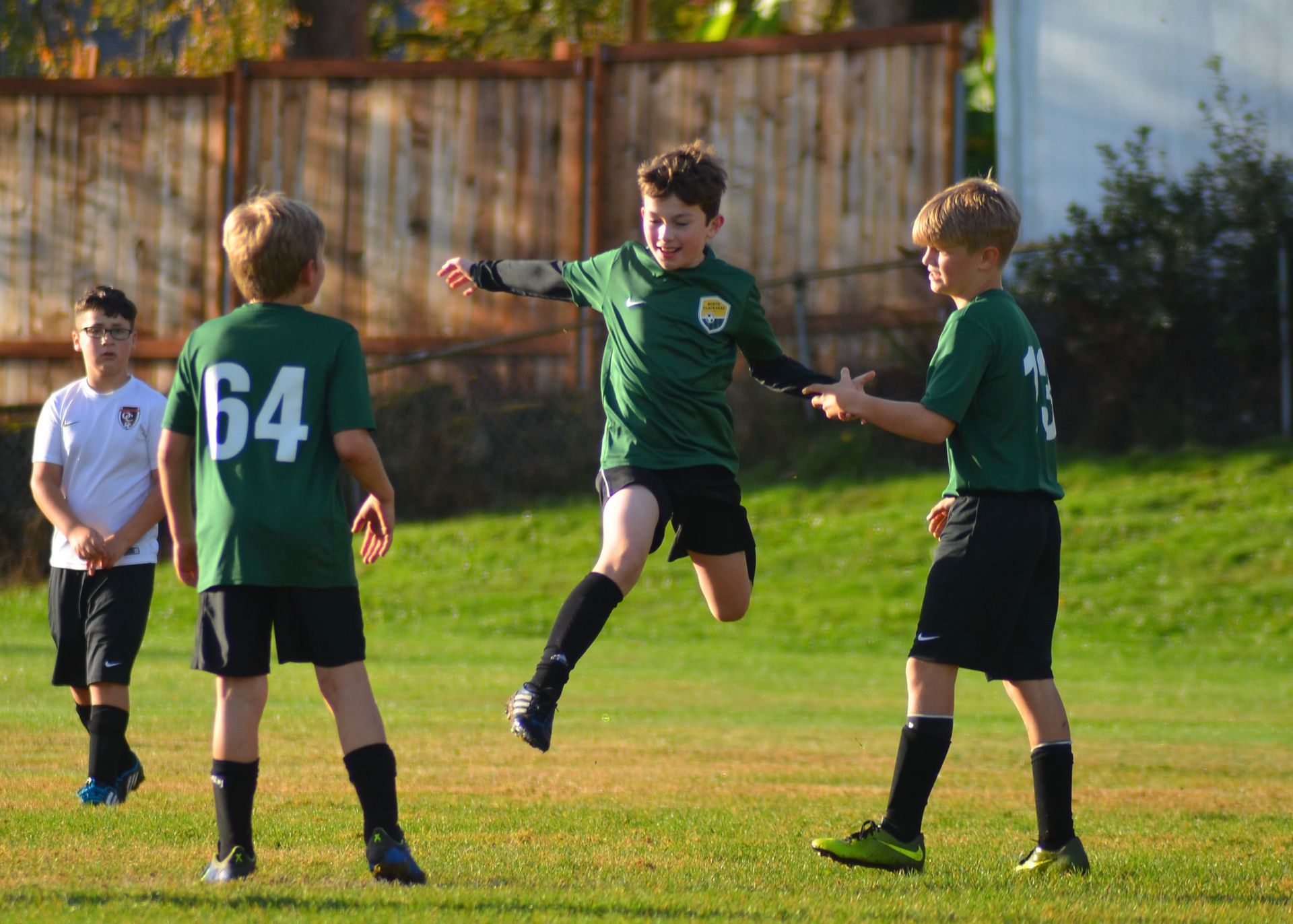 Boys in green soccer uniforms playing on a field; one is mid-air, arms out, others watch.