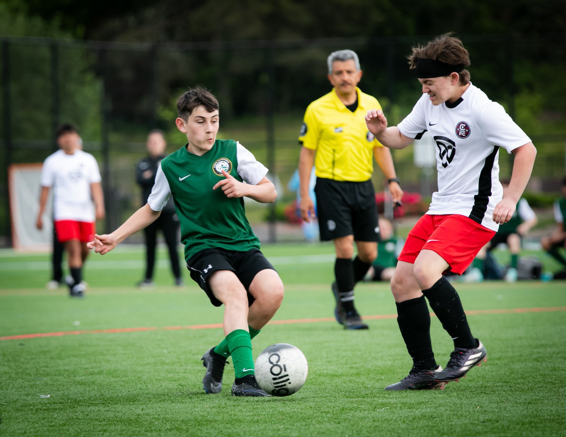 Soccer players on green field, one in green jersey dribbling ball, opponent in white, referee in yellow behind.